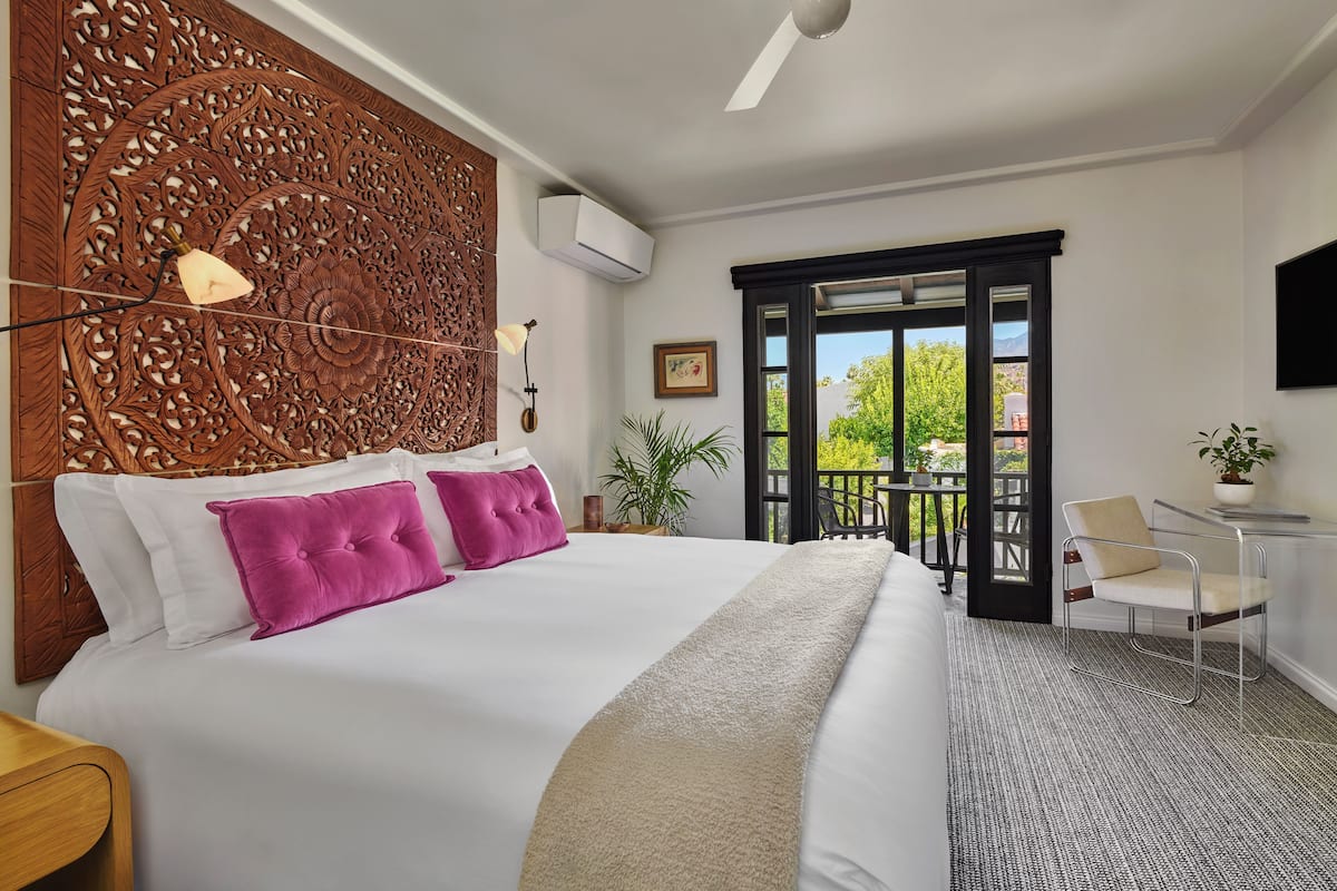 A hotel room with a large bed, a decorative headboard, a balcony with a chair, and a TV mounted on the wall.