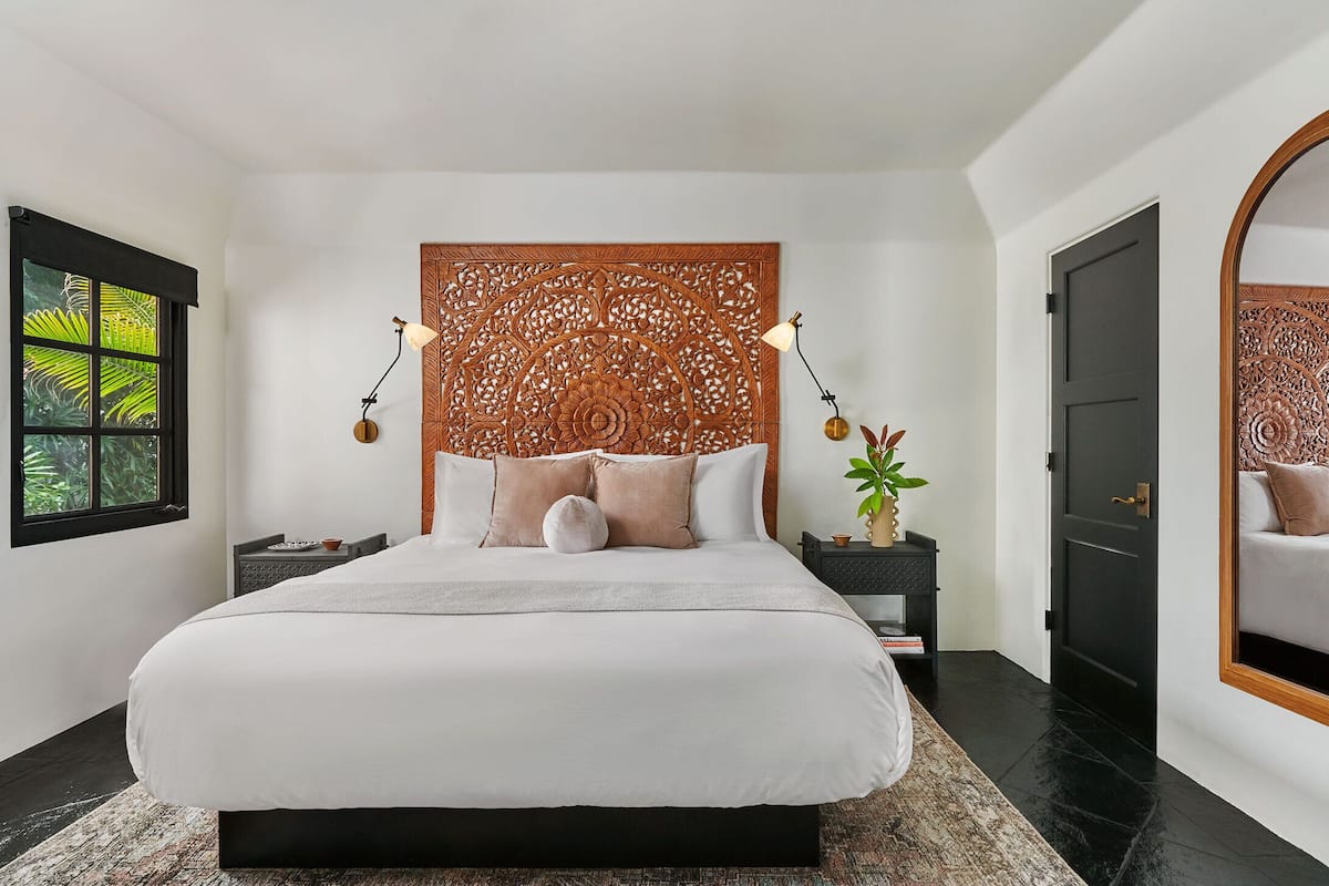A modern bedroom with a large bed, a decorative headboard, bedside tables, a mirror, and a view of greenery through a window.
