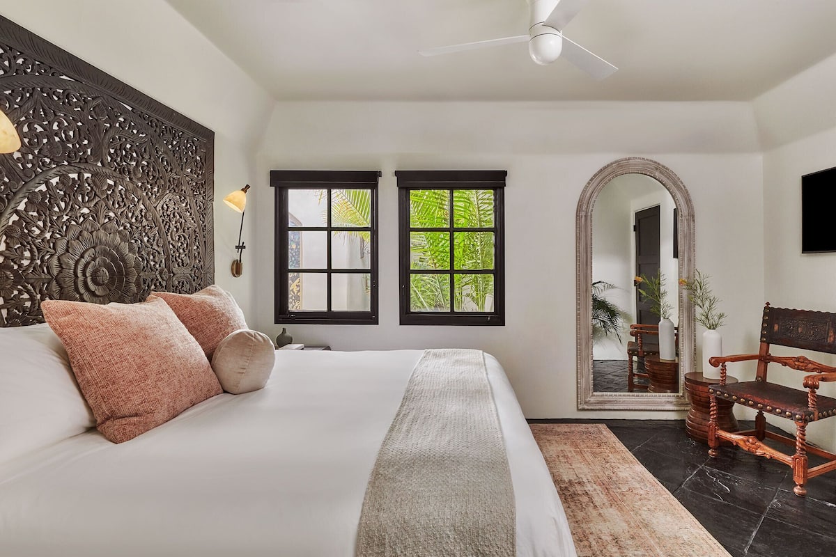 A bedroom with a large bed, two windows, a ceiling fan, a mirror, and a wooden chair.