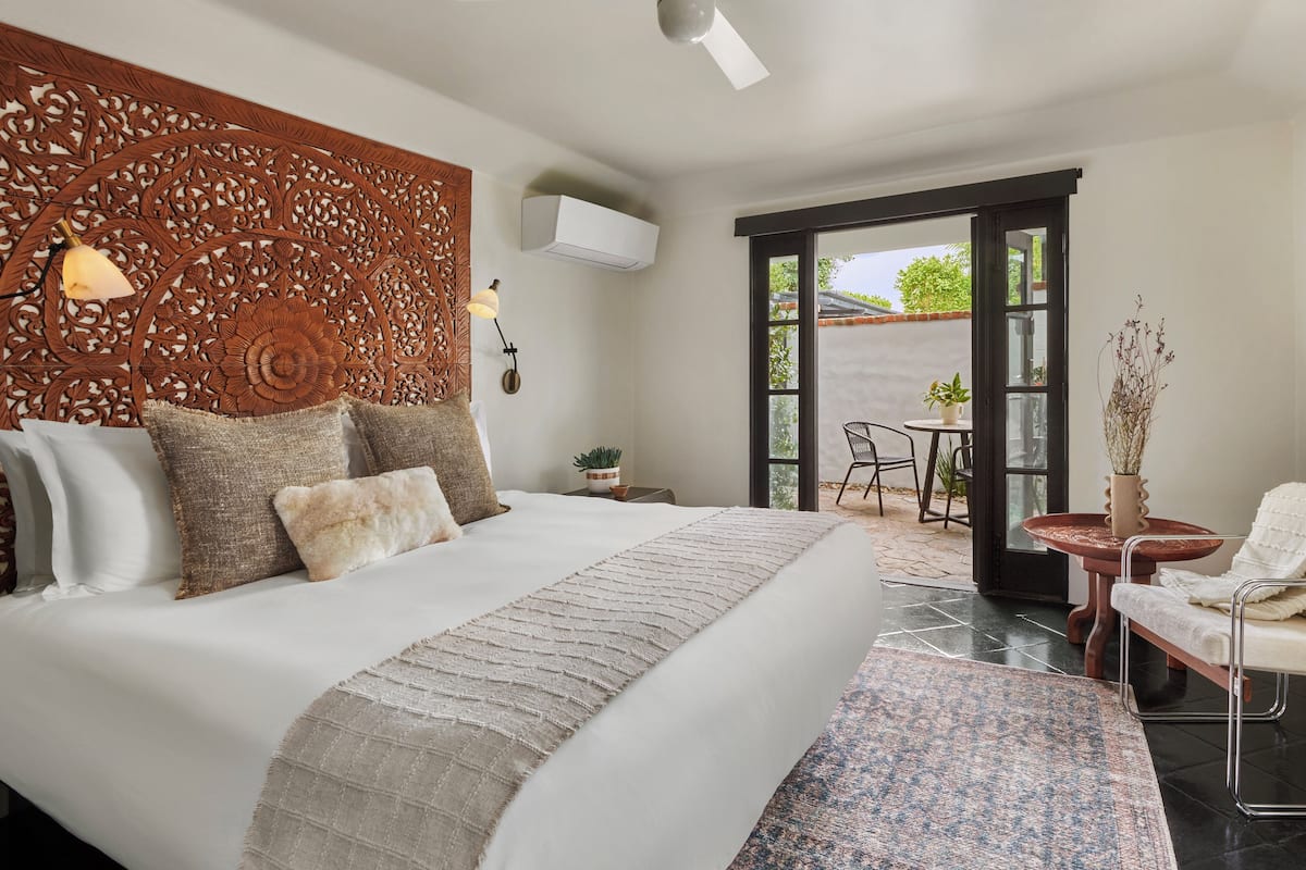 A bedroom with a large bed, a decorative headboard, an air conditioning unit, a small table with a plant, and a view of the outdoors.
