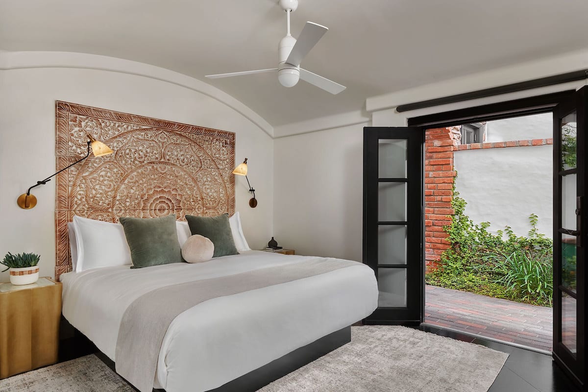 A modern bedroom with a large bed, a ceiling fan, and a view of a brick wall and greenery outside.