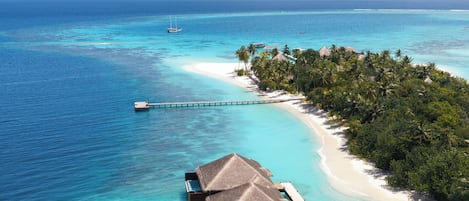An overwater bungalows with thatched roofs, connected by a wooden walkway, overlooking a clear turquoise sea and a sandy beach with palm trees.