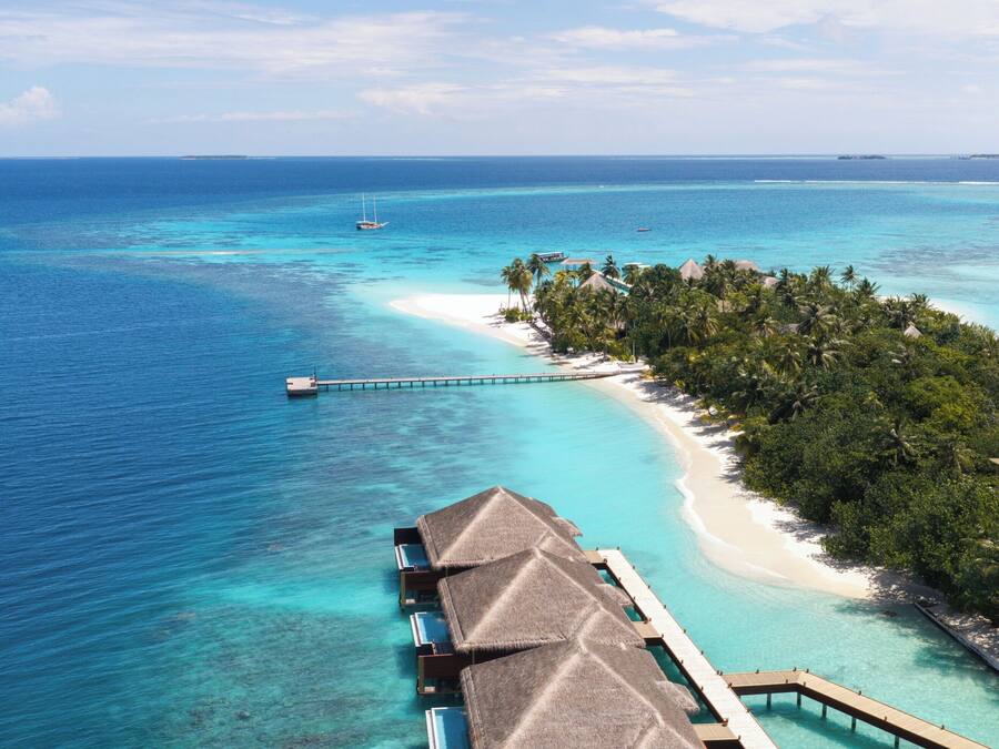 An overwater bungalows with thatched roofs, connected by a wooden walkway, overlooking a clear turquoise sea and a sandy beach with palm trees.