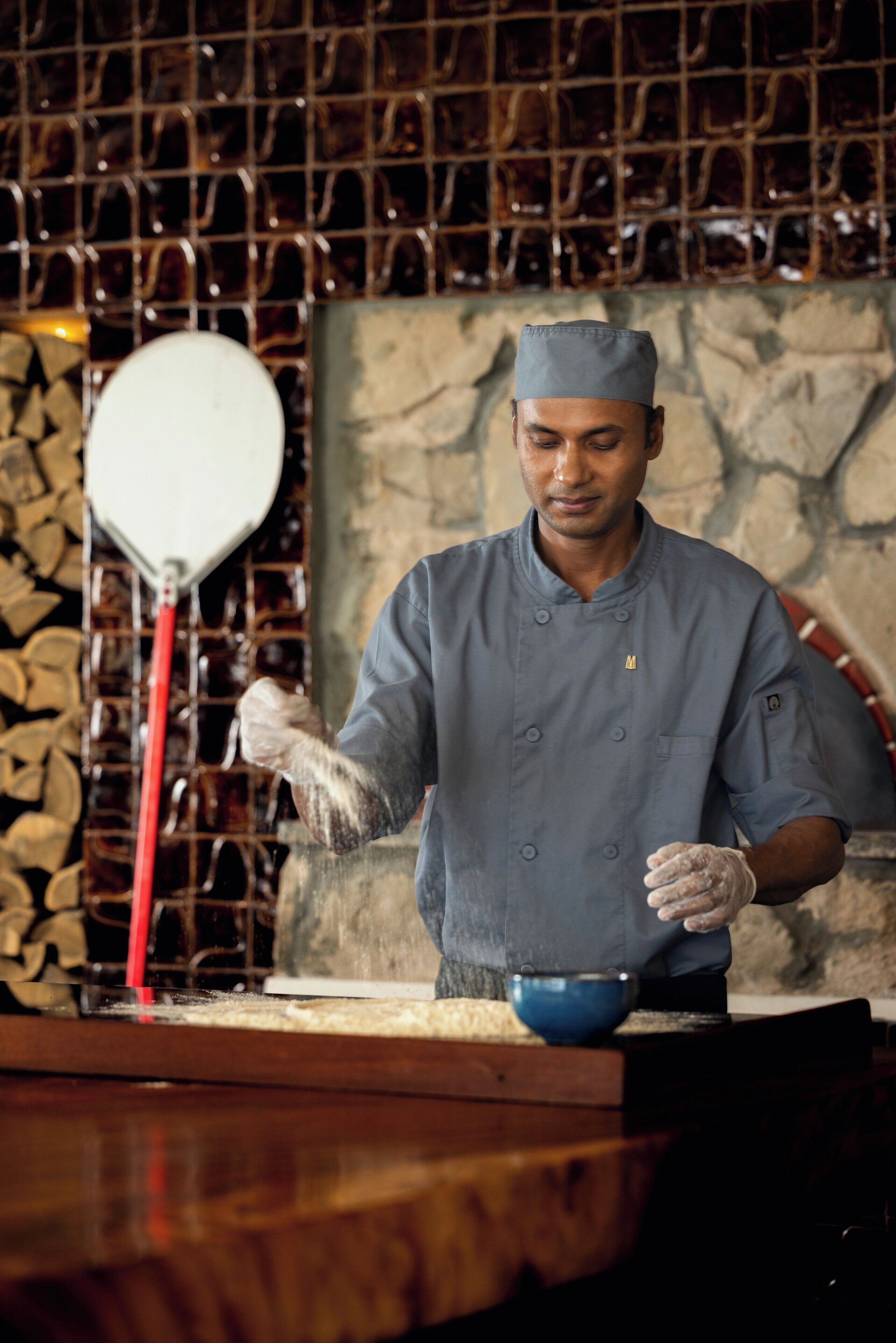 A chef in a uniform with a cap, standing in a kitchen with a stone wall and a rack of glasses in the background.