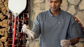 A chef in a uniform with a cap, standing in a kitchen with a stone wall and a rack of glasses in the background.