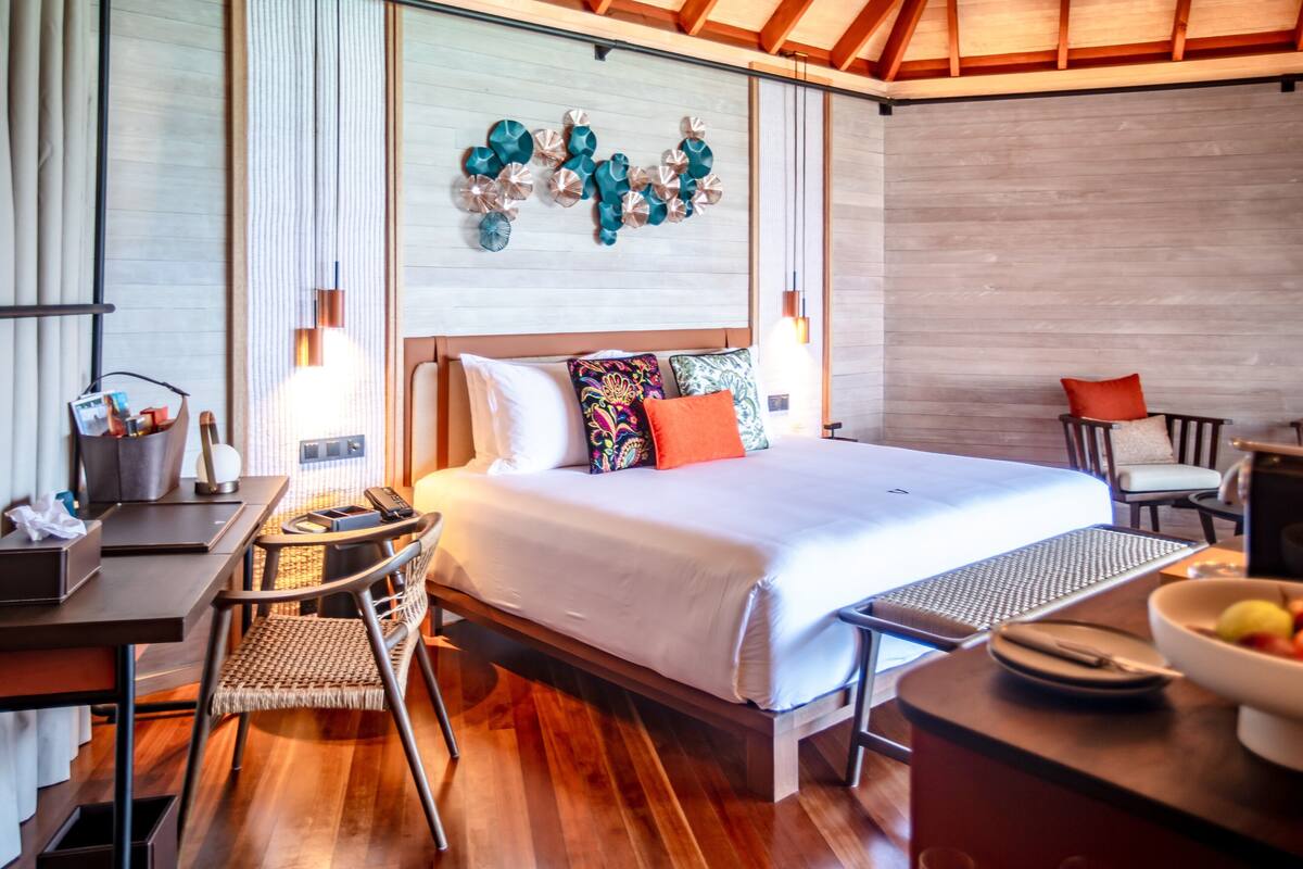 A modern hotel room with a wooden floor, a bed with white bedding, a desk with a chair, and a wall-mounted light fixture.