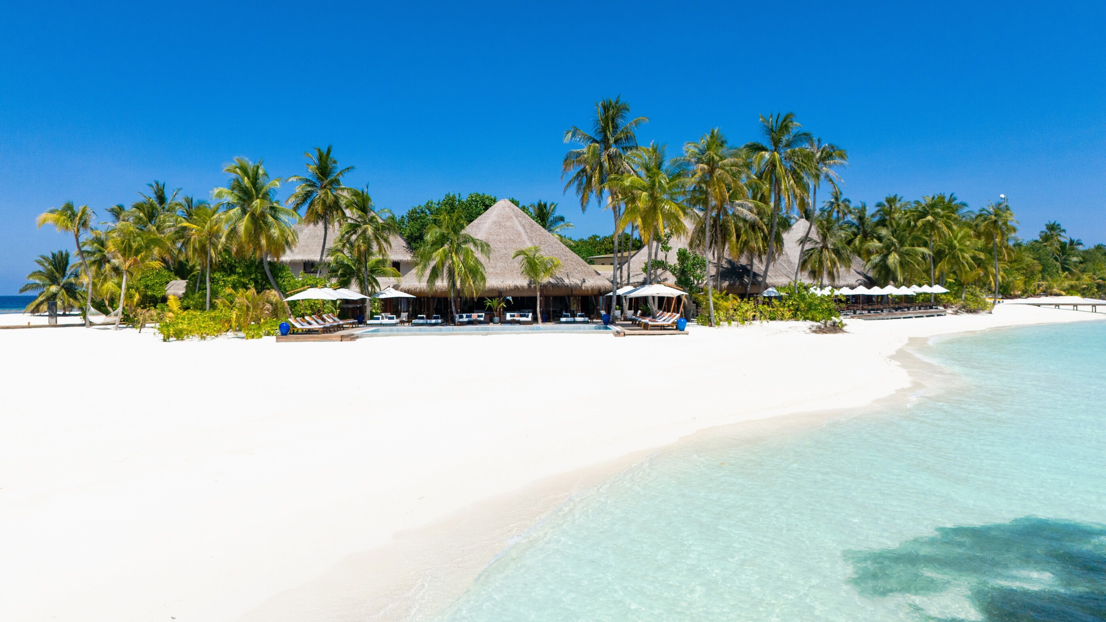 A beach resort with thatched-roof huts, palm trees, and a clear blue sky.