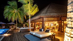 Una terraza de madera con piscina, zona de descanso y construcción de techo de paja que muestra espacios interiores para comer y sentarse.