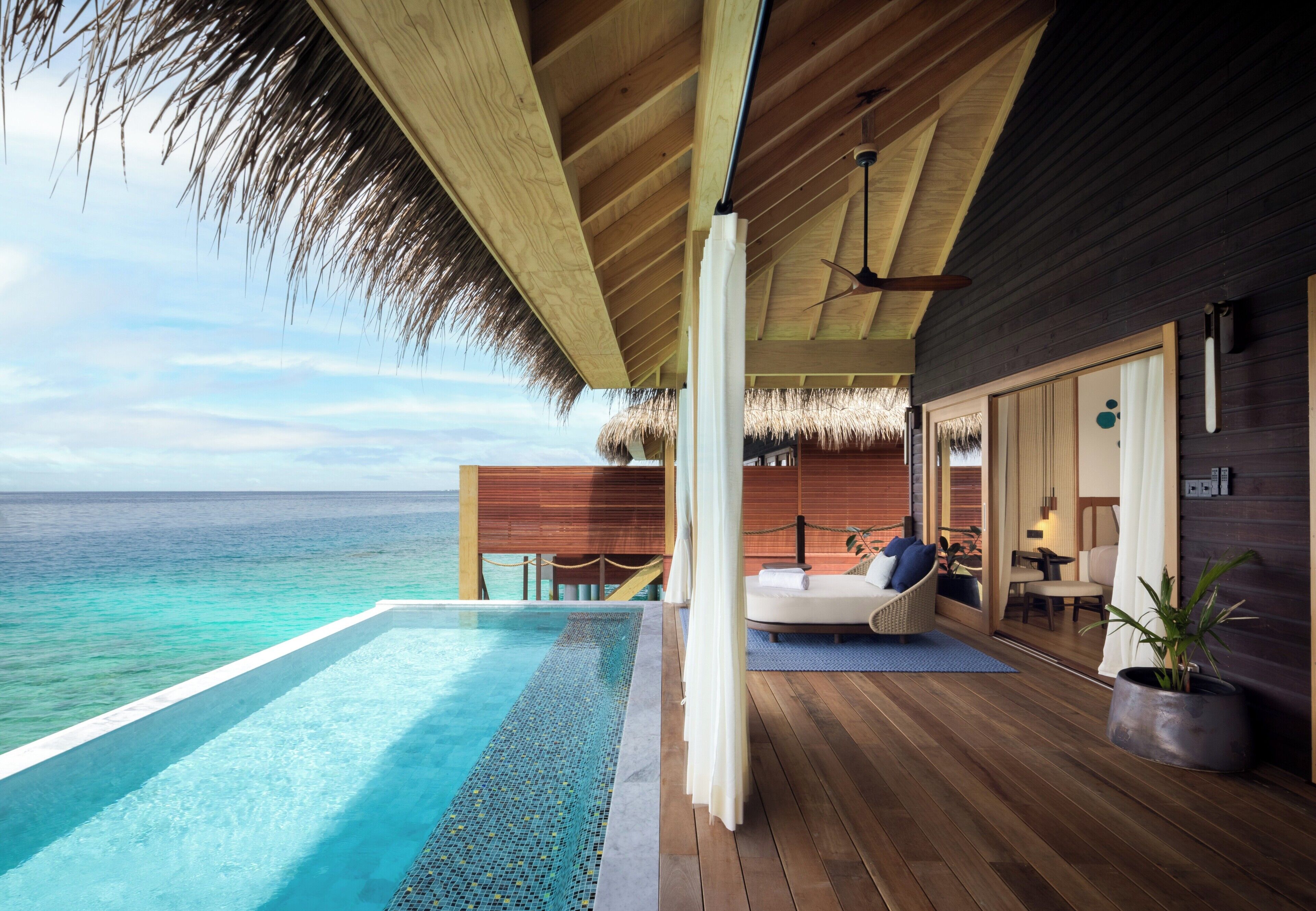 An infinity pool with a clear view of the ocean, a wooden deck, a thatched roof, and a modern interior with a bed and seating area.
