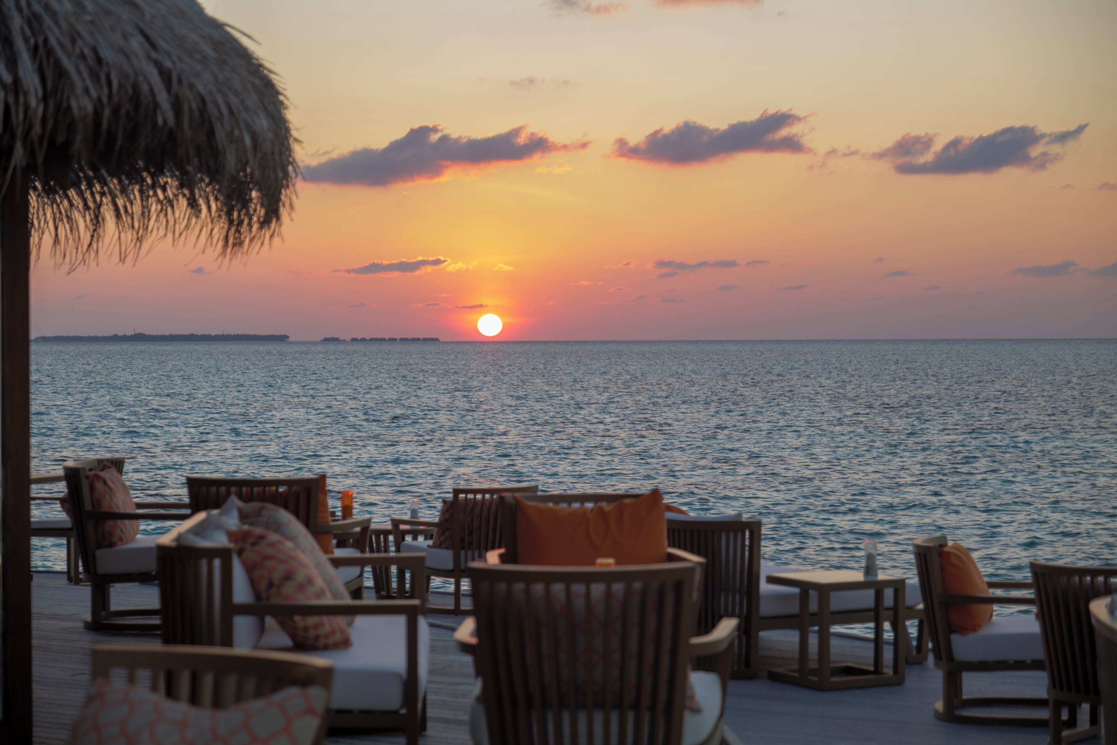 A sunset view from a deck with outdoor seating and a thatched roof.