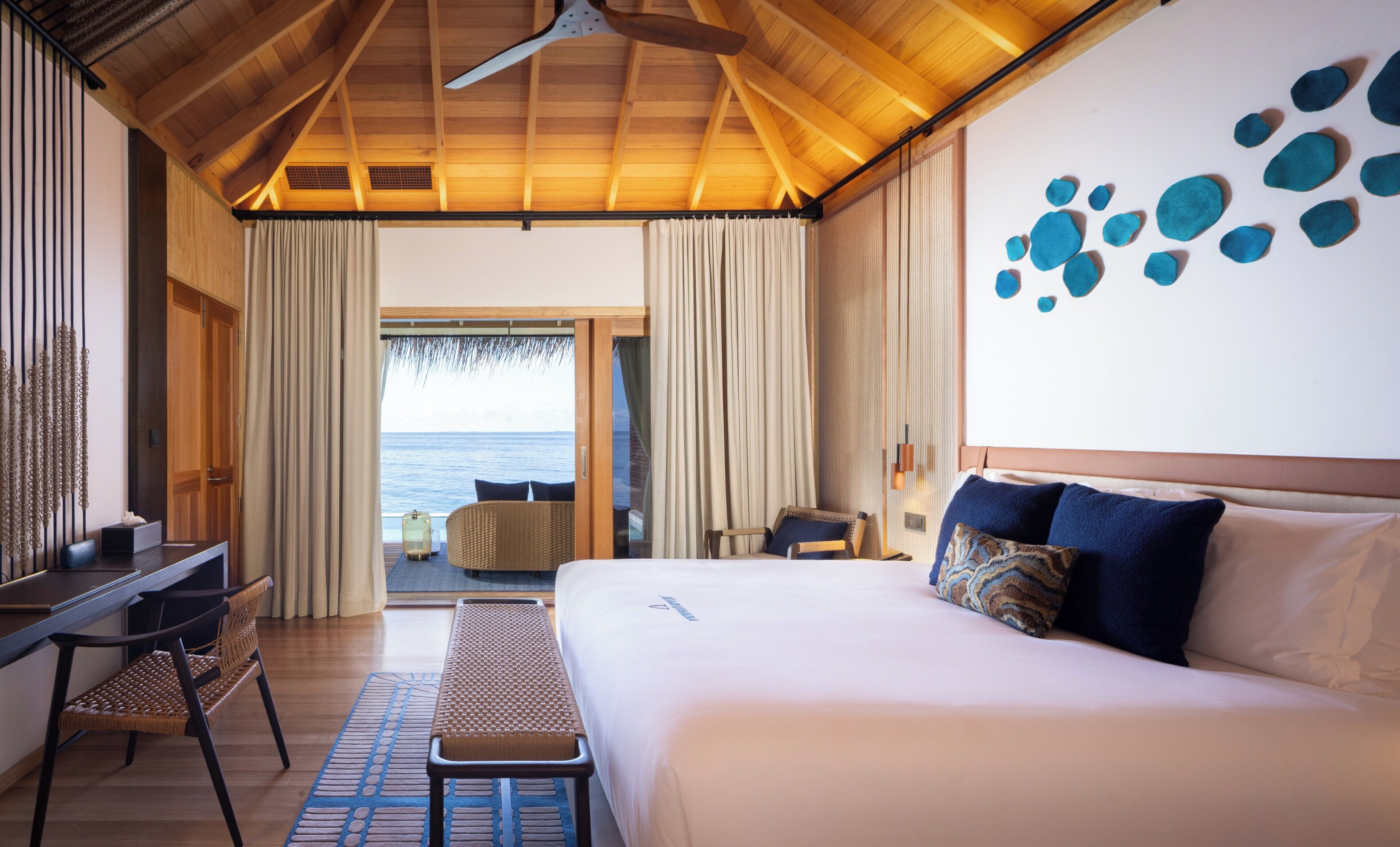 Quarto de hotel com cama, escrivaninha, cadeira, um banco e vista para o oceano.
