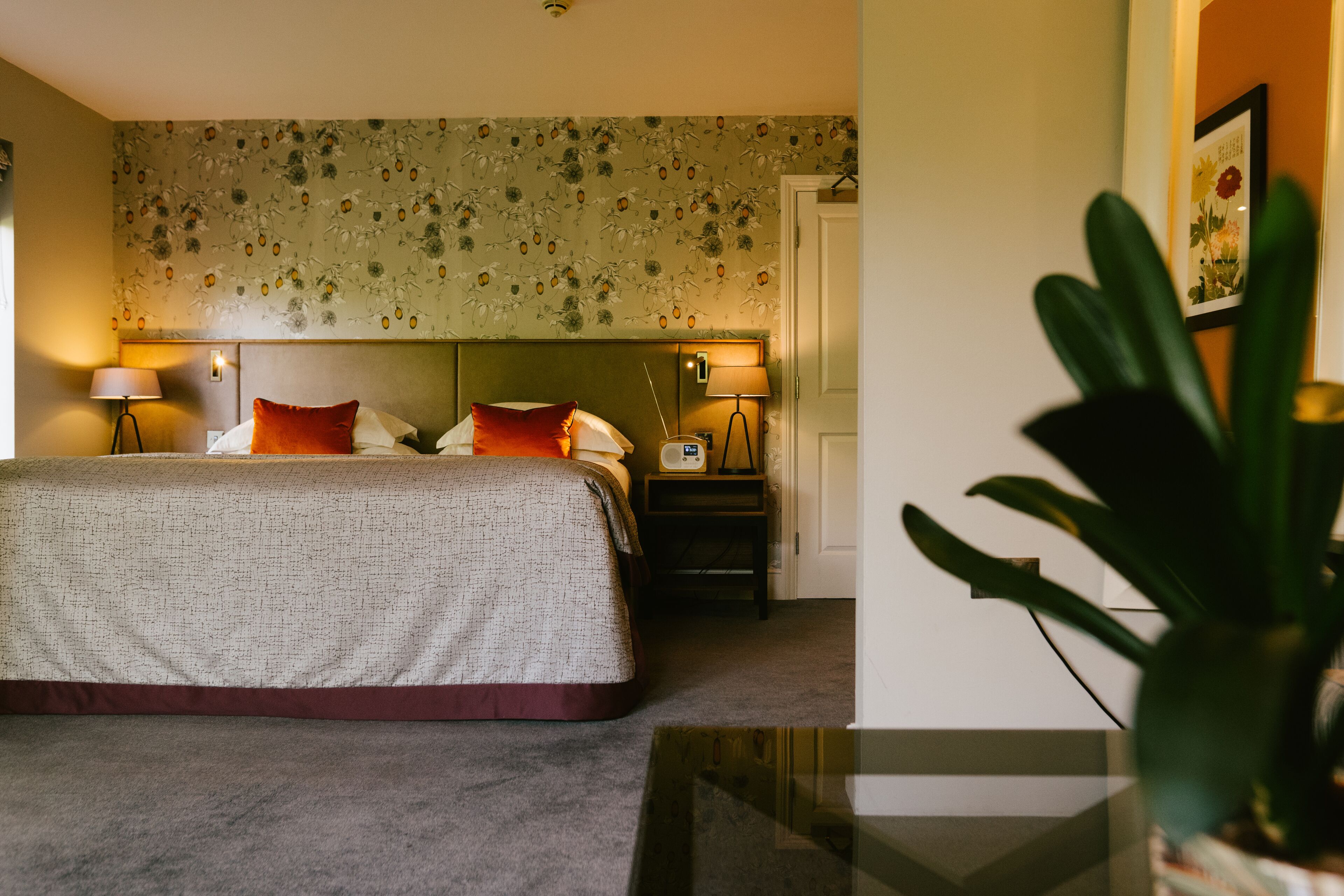 Una habitación de hotel con una cama grande, dos mesitas de noche con lámparas, papel tapiz con motivos florales y una planta en maceta.