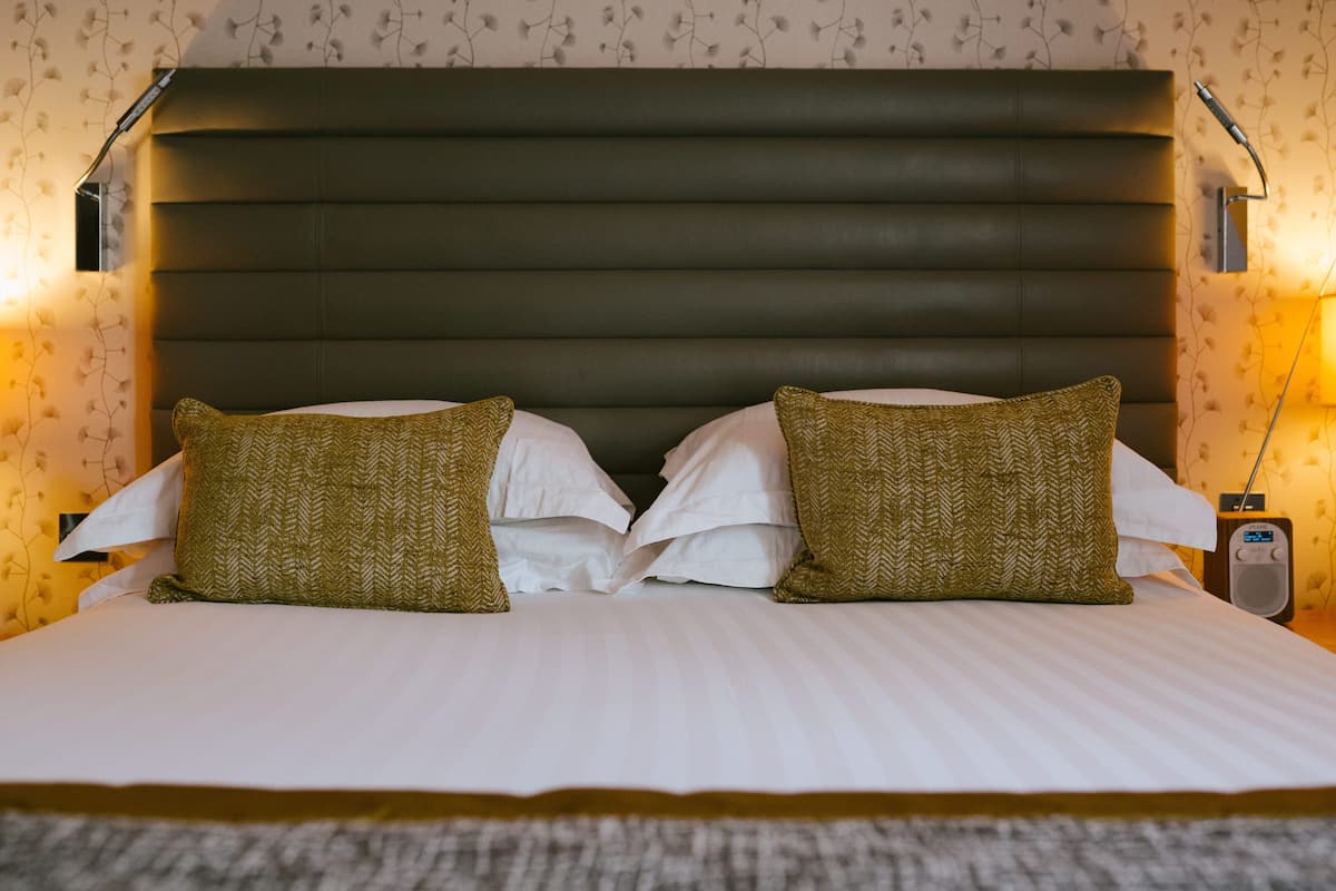 Una cama bien hecha con un cabecero oscuro, dos almohadas y una mesita de noche con una lámpara y una radio.