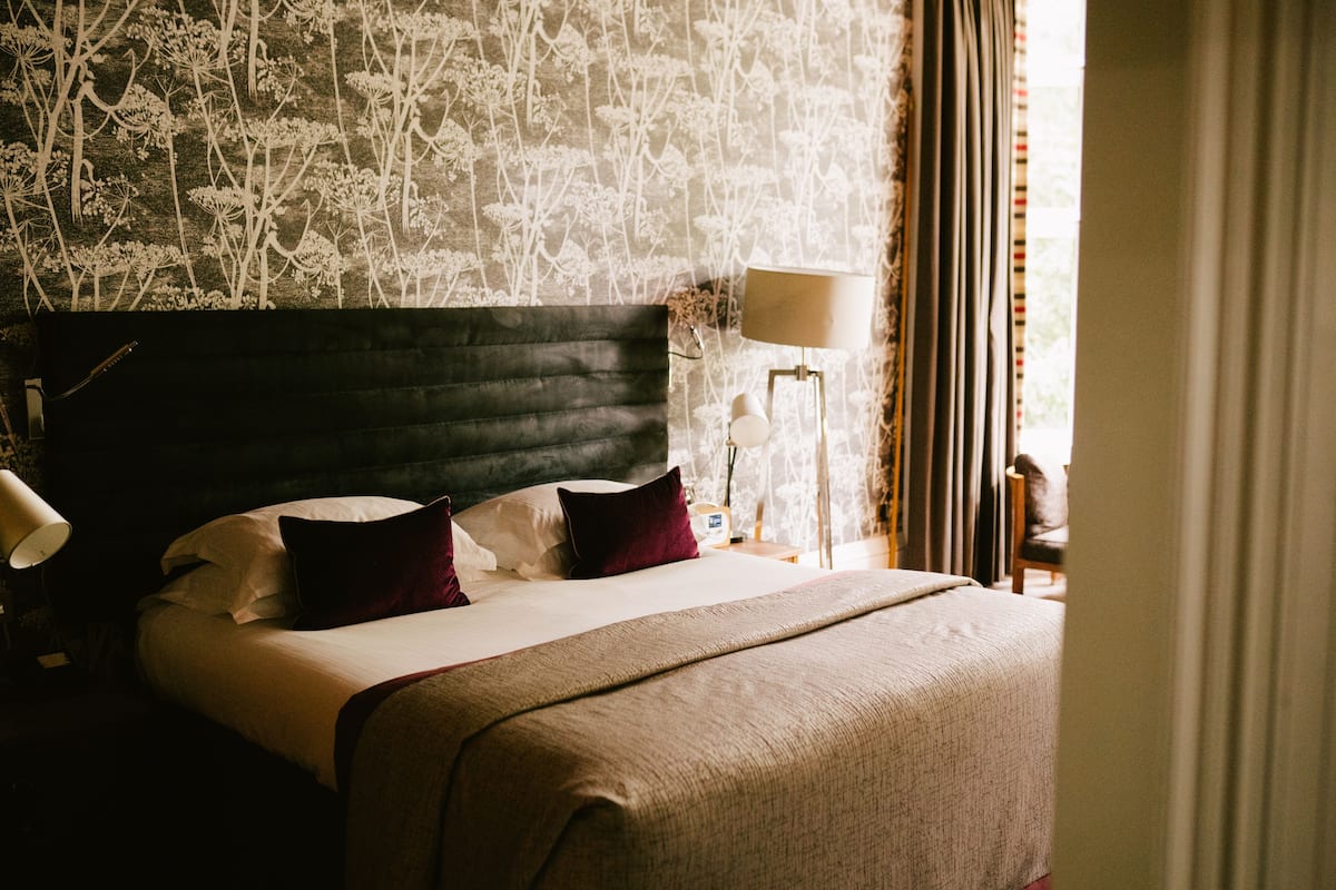 Una cama bien hecha con un cabecero oscuro, almohadas blancas y rojas y una manta color beige. Hay una lámpara de noche y una mesita con una lámpara y un objeto decorativo. La pared tiene un estampado de papel tapiz floral.