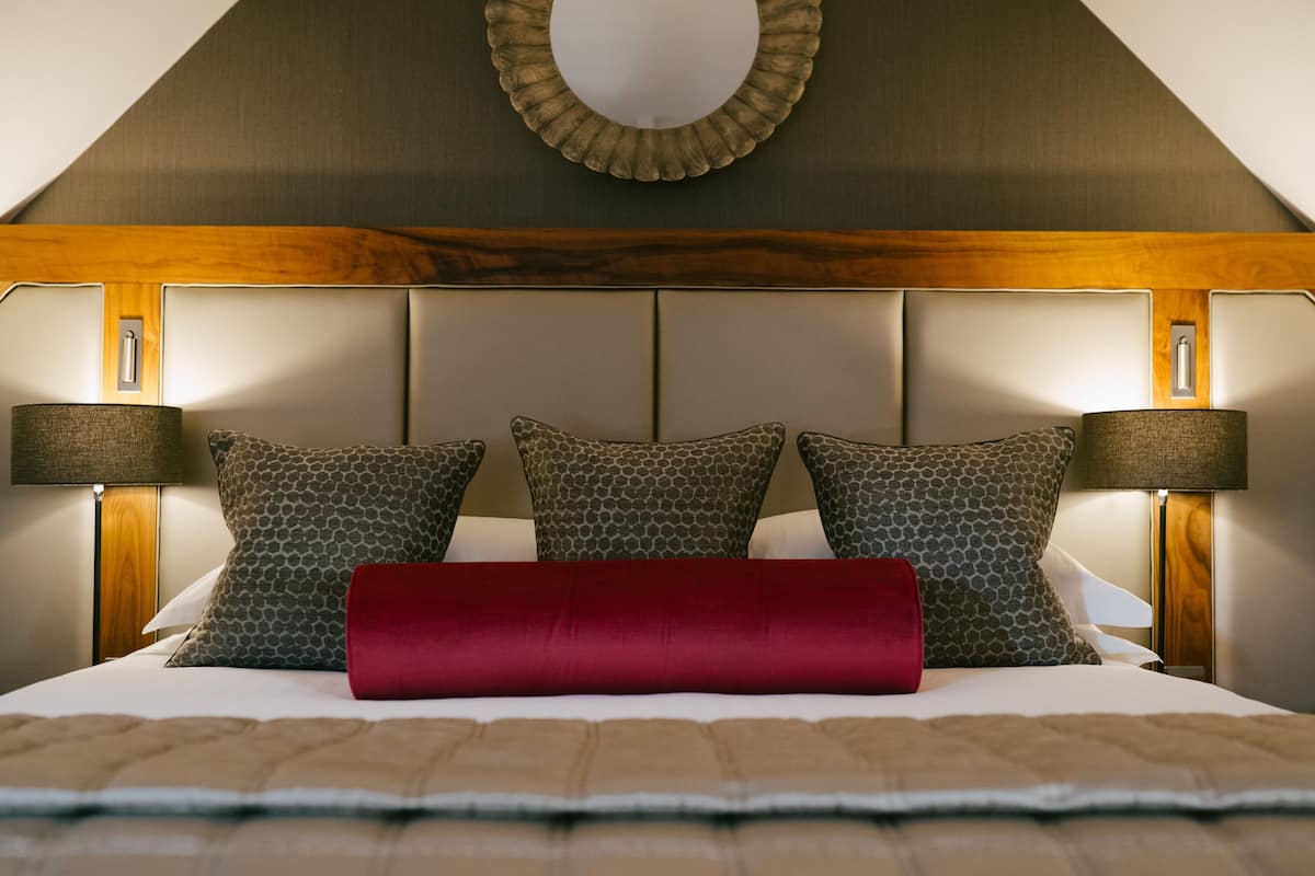 Una cama bien tendida con almohadas estampadas y una almohada roja decorativa, flanqueada por dos lámparas de noche, en una habitación con cabecera de madera y un espejo circular arriba.