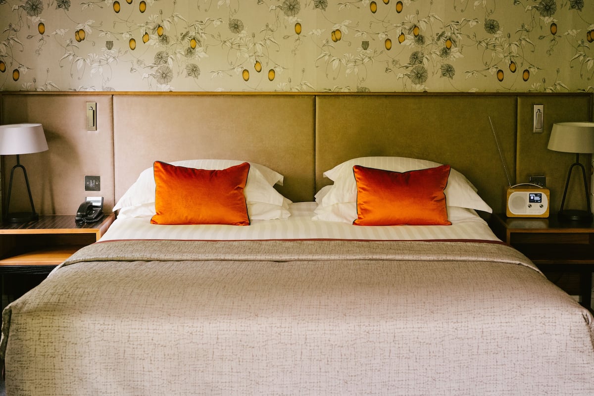 Una habitación de hotel con cama, dos almohadas, mesita de noche, lámpara y radio.