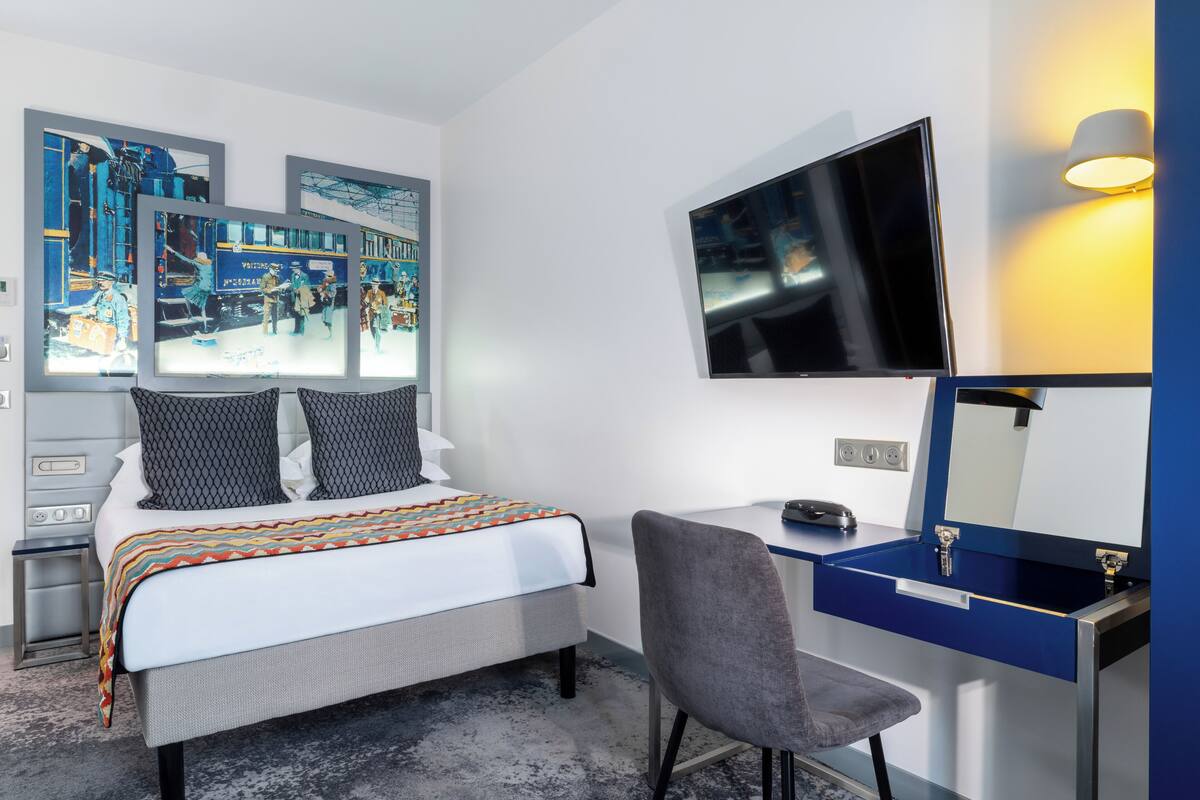 A hotel room with a bed, a desk, a chair, a TV, and artwork on the walls.