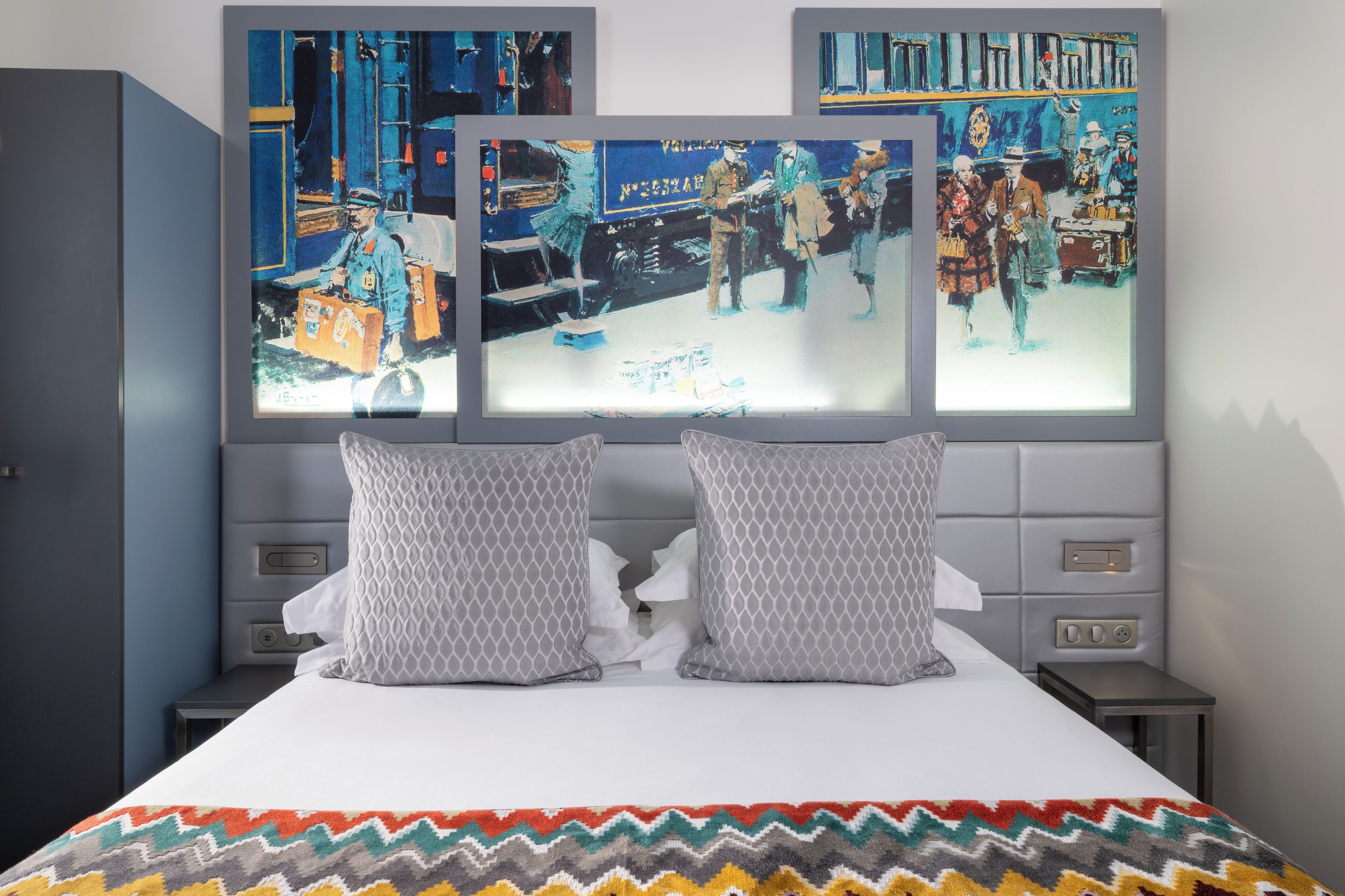 A hotel room with a bed, two gray patterned pillows, and three framed paintings on the wall.