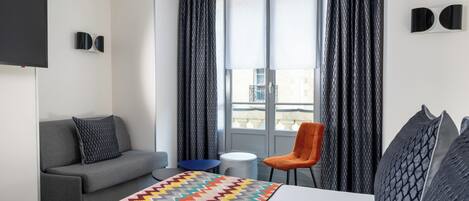 Quarto de hotel com cama, sofá cinzento, poltrona laranja e uma pequena mesa redonda.
