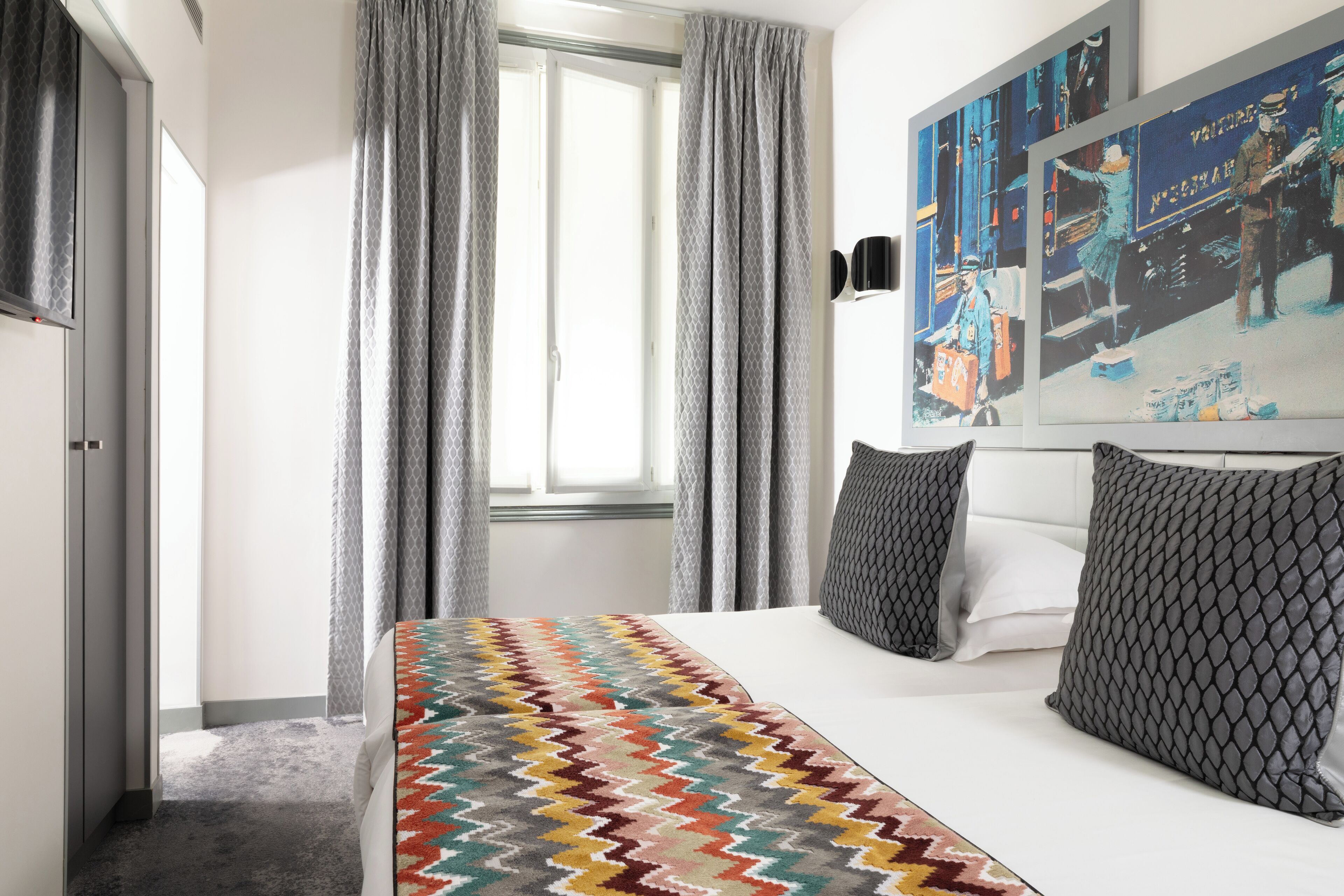 Habitación de hotel con cama, cortinas grises, colcha estampada y una pared con obras de arte.