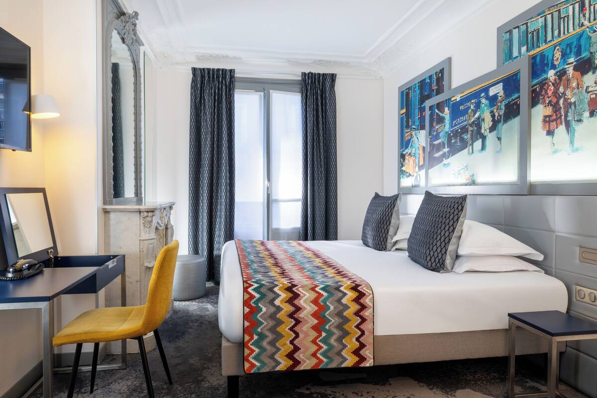 A hotel room with a bed, a desk, a chair, and a large window with curtains.