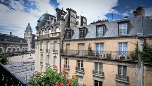 Un edificio de varios pisos con balcones, una torre de reloj y vista a una calle concurrida con coches y peatones.
