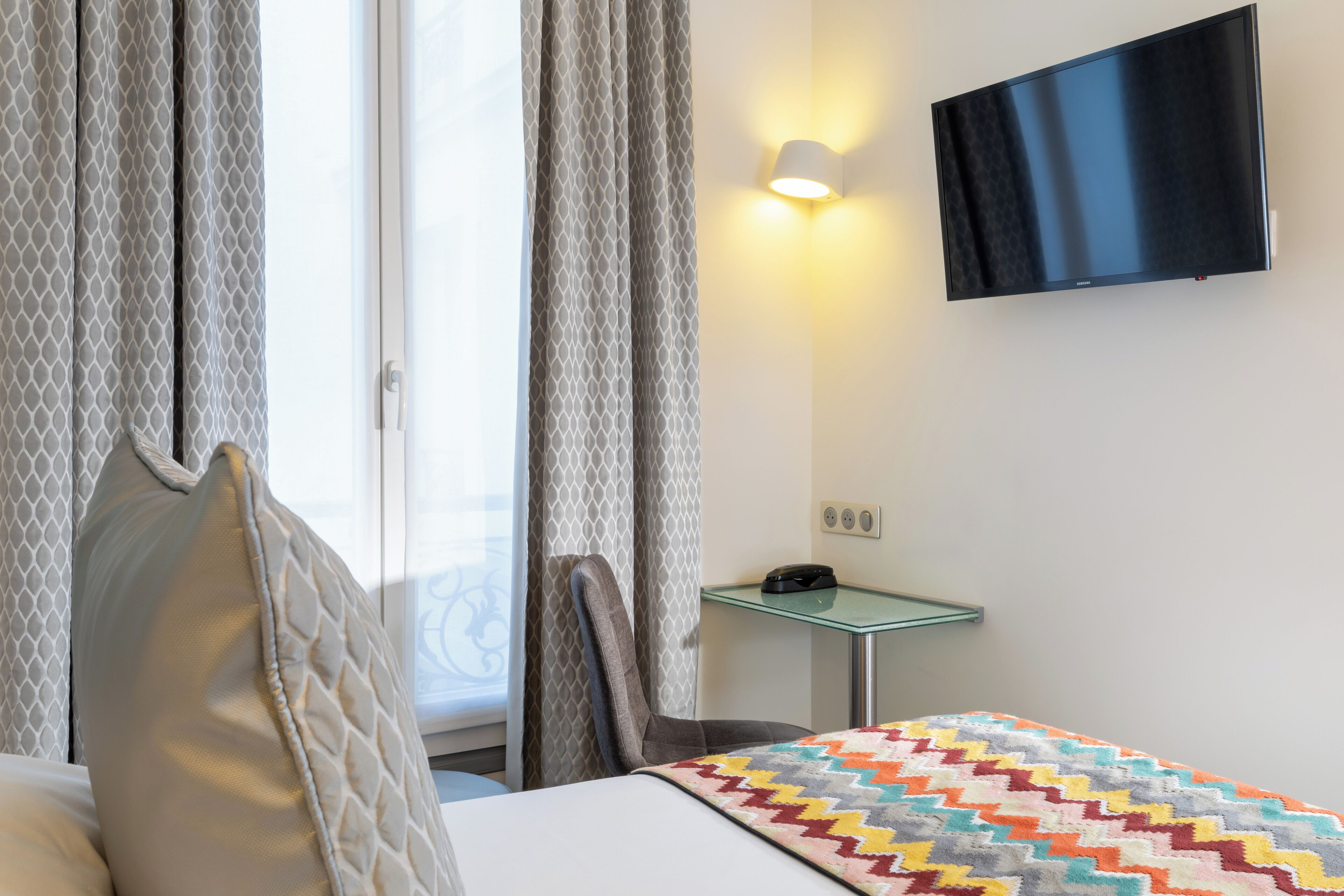 A hotel room with a bed, a TV mounted on the wall, a small table with a phone, and a window with curtains.