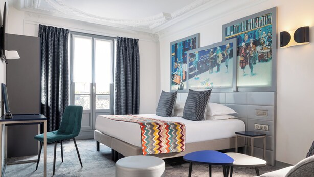 Una habitación de hotel con una cama grande, un escritorio, una silla, una mesita y un sofá. Hay cortinas en las ventanas y obras de arte en las paredes.