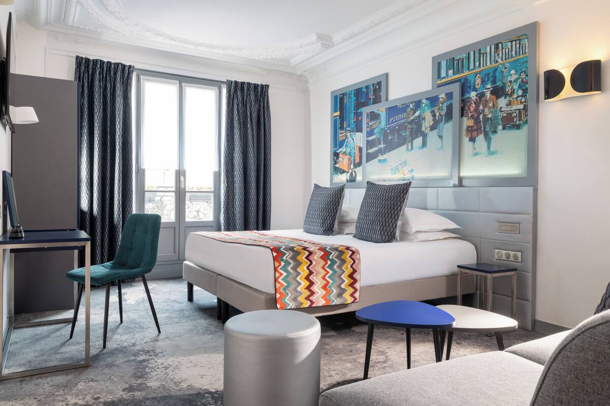 A hotel room with a large bed, a desk, a chair, a small table, and a sofa. There are curtains on the windows and artwork on the walls.
