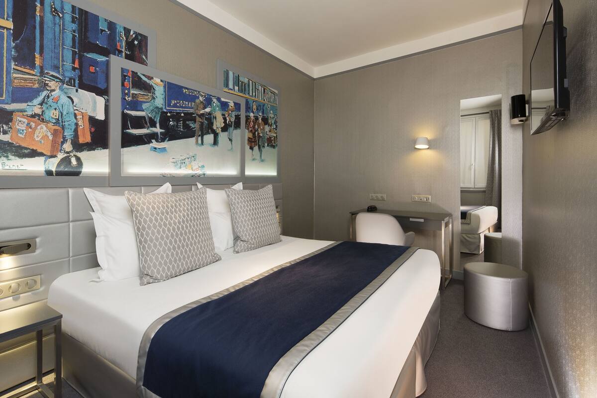 A hotel room with a bed, a desk, a chair, a mirror, and artwork on the wall.