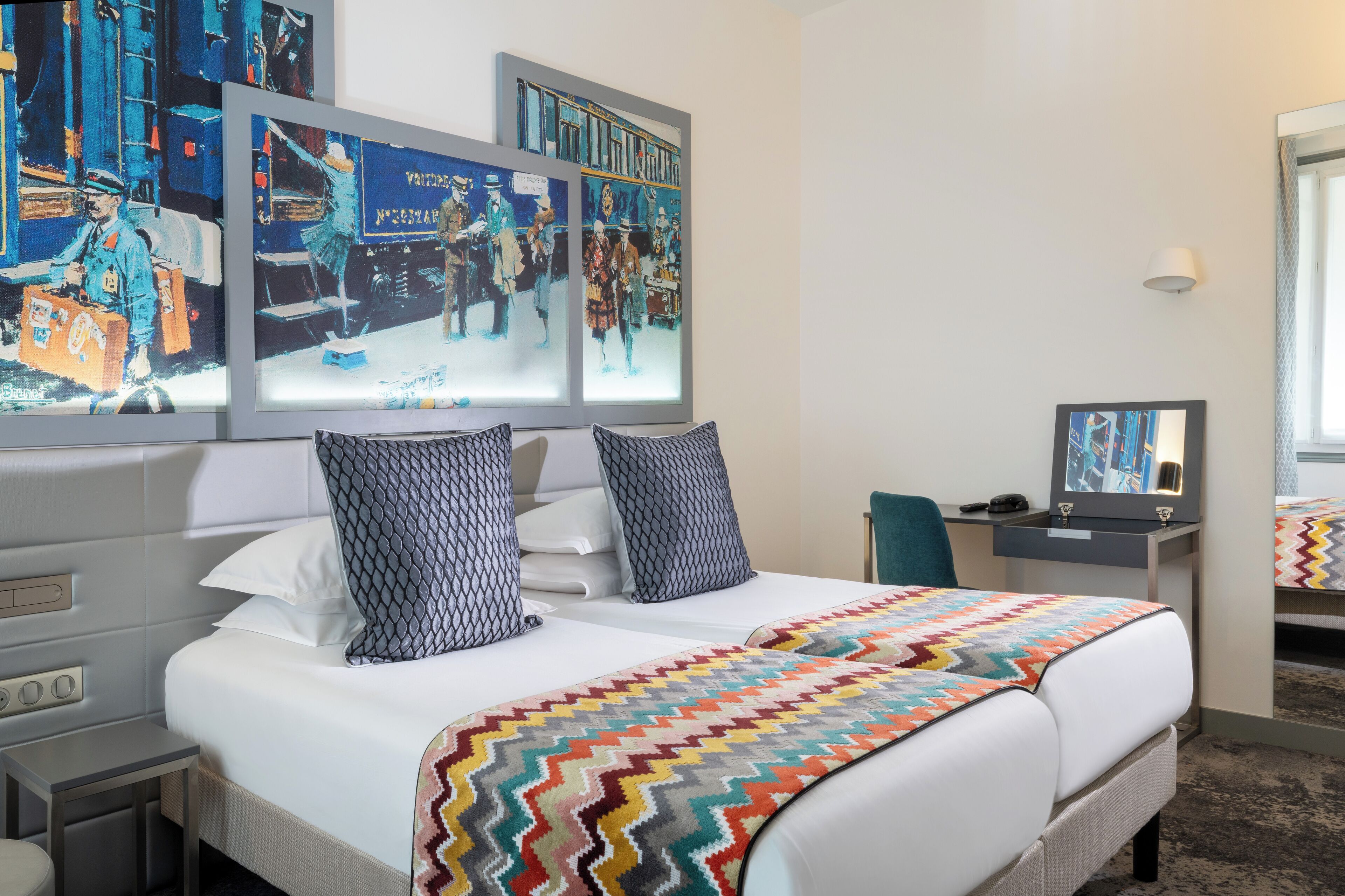 Ein Hotelzimmer mit einem großen Bett, zwei gemusterten Kissen, einer farbenfrohen Decke, einem Schreibtisch mit einer Lampe und Bildern an der Wand.