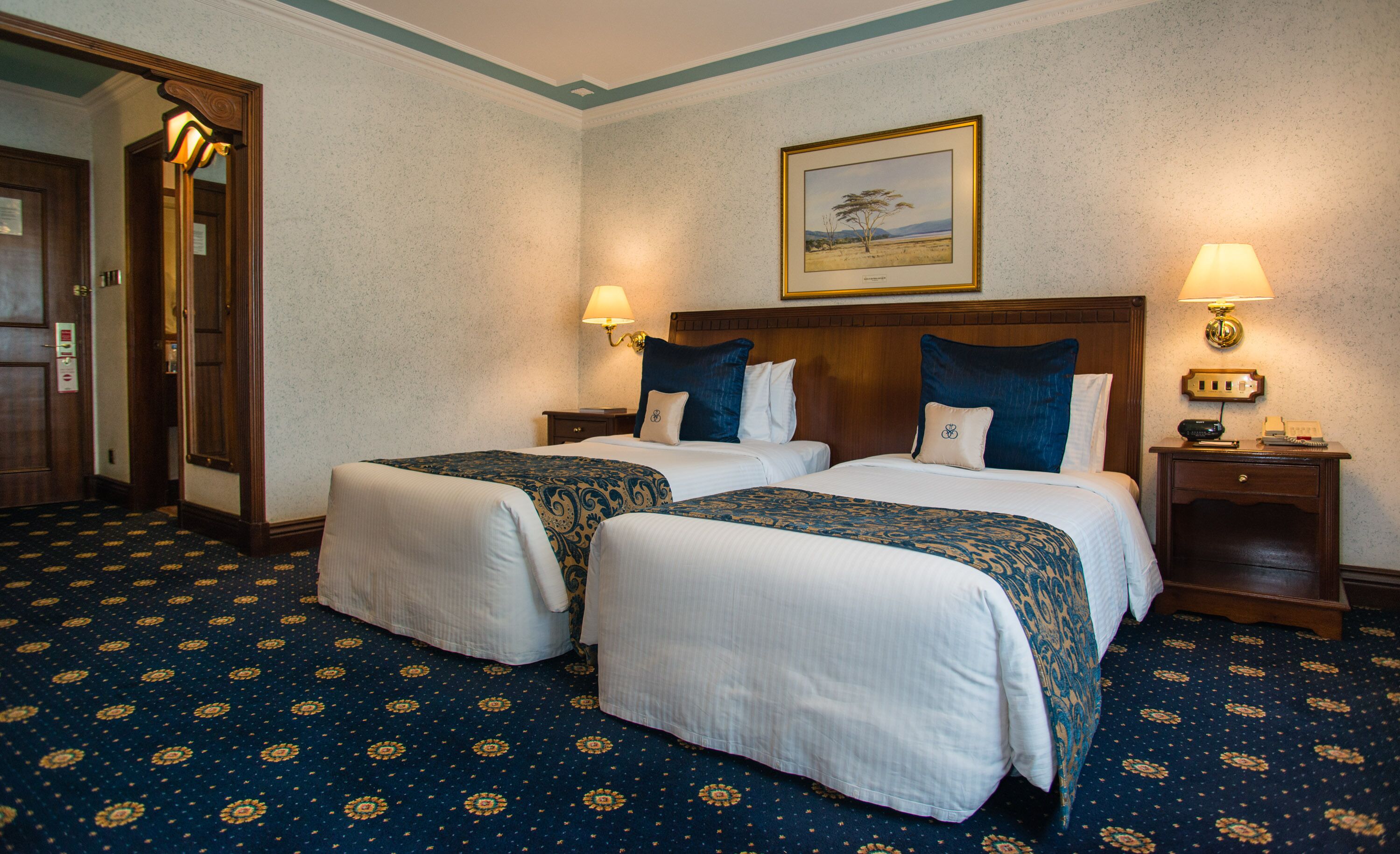 A hotel room with two beds, a large mirror, a painting on the wall, and a carpet with a floral pattern.