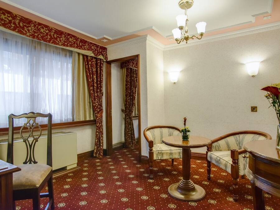 Una habitación con alfombra estampada, una mesa de comedor con sillas, un radiador y una ventana con cortinas.