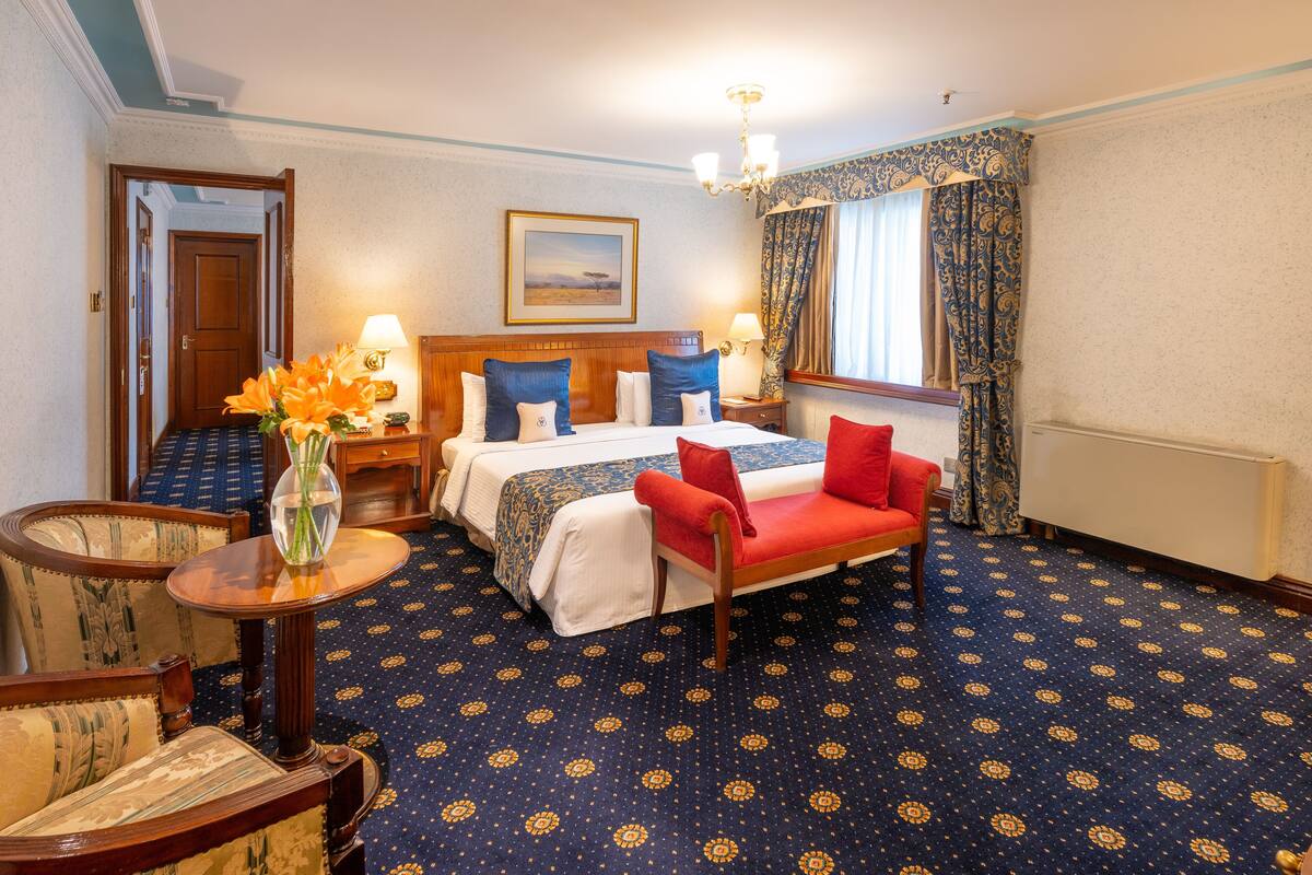 A hotel room with a large bed, a red armchair, a wooden nightstand, a table with a vase of flowers, and a window with curtains.