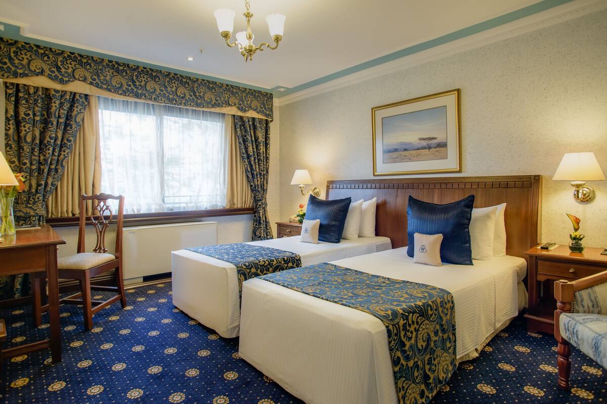 A hotel room with two beds, a large window with curtains, a chandelier, and a painting on the wall.