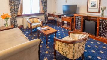 Hotellihuone, jossa on sohva, kaksi nojatuolia, pieni pöytä, televisio, kaappi ja kuviollinen matto.