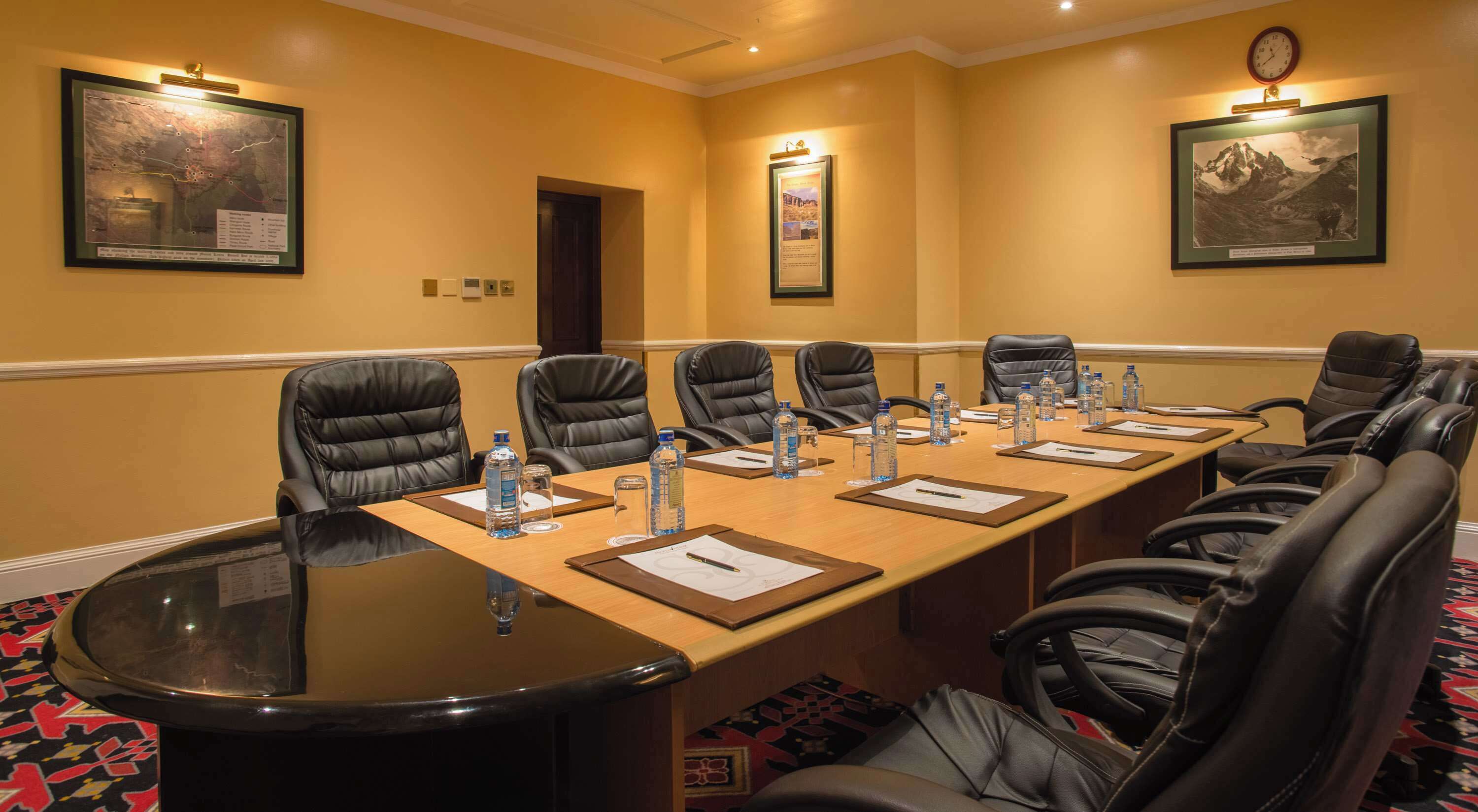 Un salón de conferencias con una larga mesa, sillas de cuero negro, botellas de agua y cuadros en las paredes.