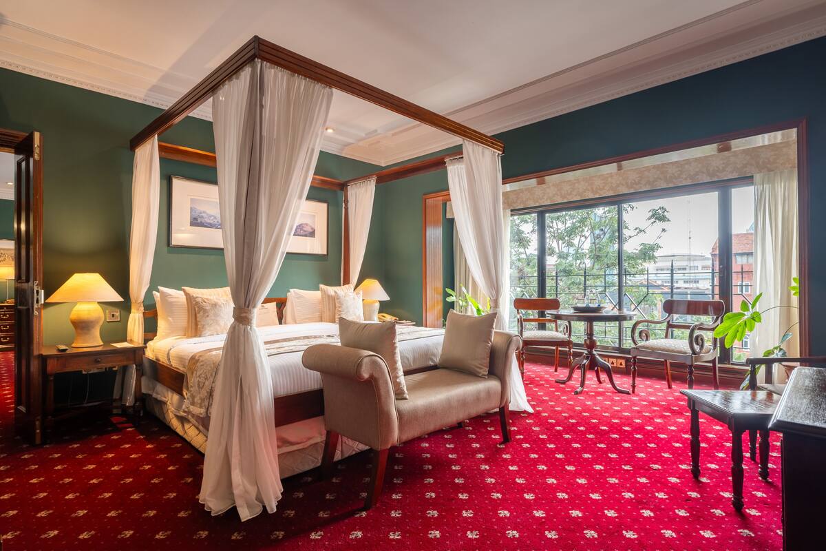 A four-poster canopy bed with a red carpeted floor, a sitting chair, and a table with chairs.