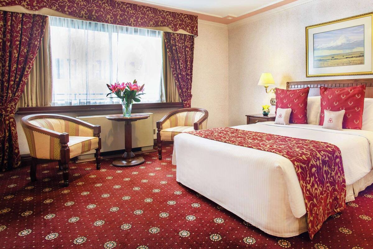 A hotel room with a large bed, two bedside tables with lamps, a round table with a vase of flowers, two armchairs, and a window with curtains.