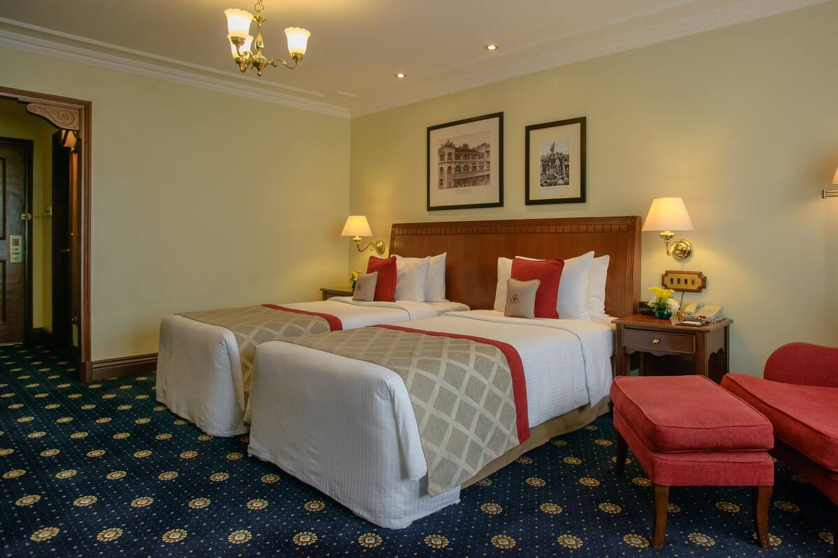 A hotel room with two beds, a red armchair, a nightstand with a lamp, and a framed picture on the wall.