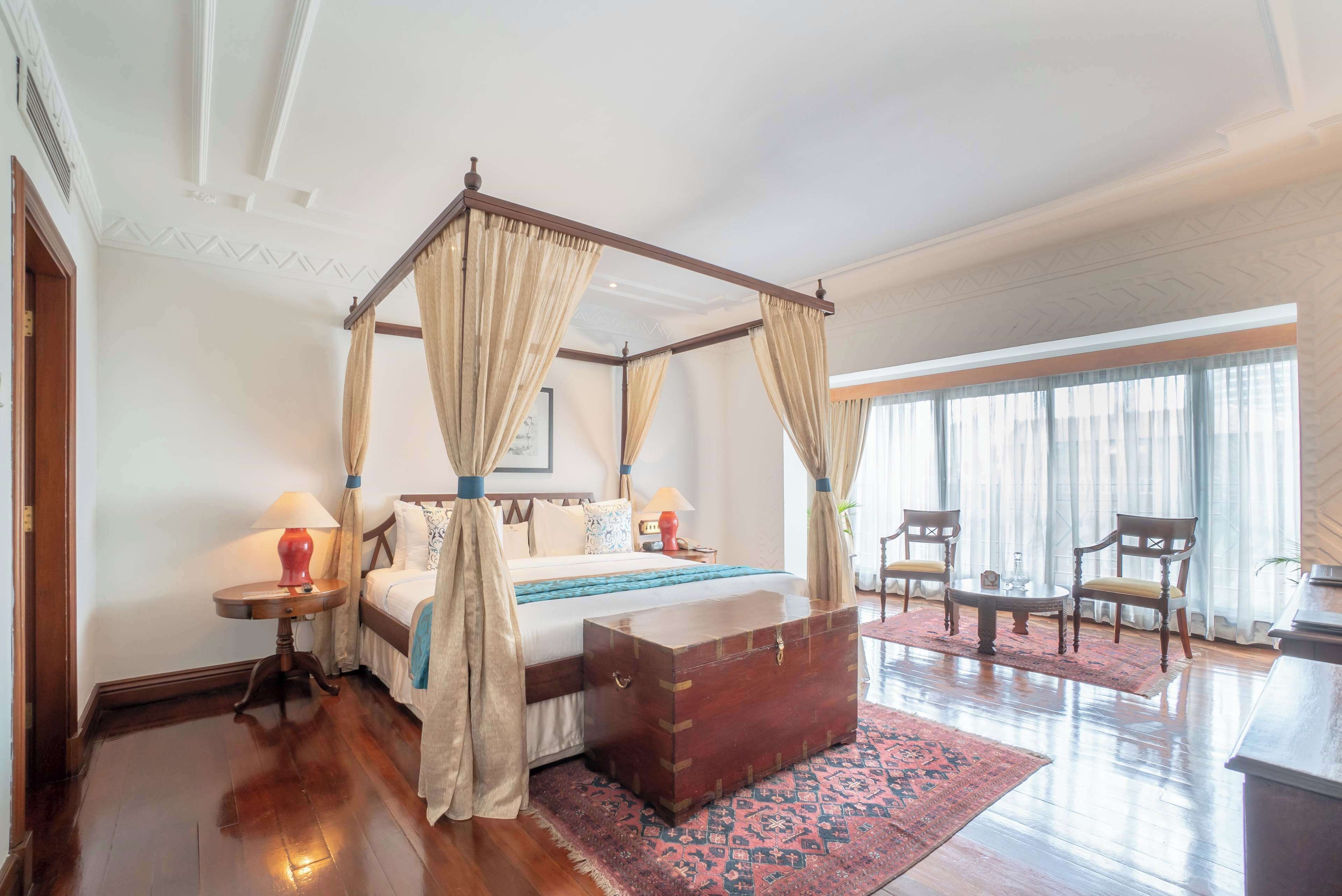 A four-poster bed with a wooden headboard, a bedside table with a lamp, and a nightstand. There is a large window with sheer curtains and a view of the outdoors.