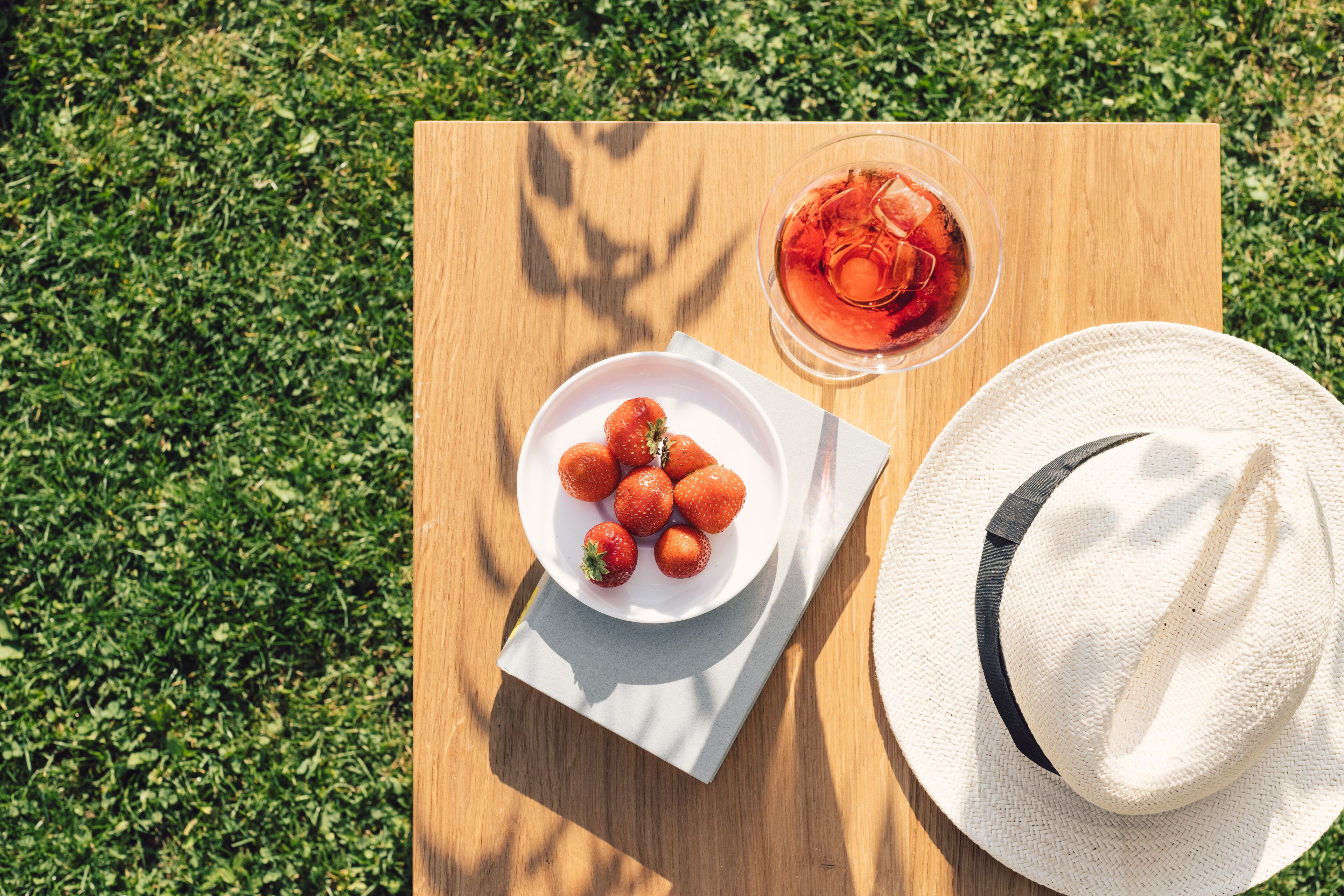 A wooden table with a white plate of strawberries, a glass of red drink, and a white hat with a black band.