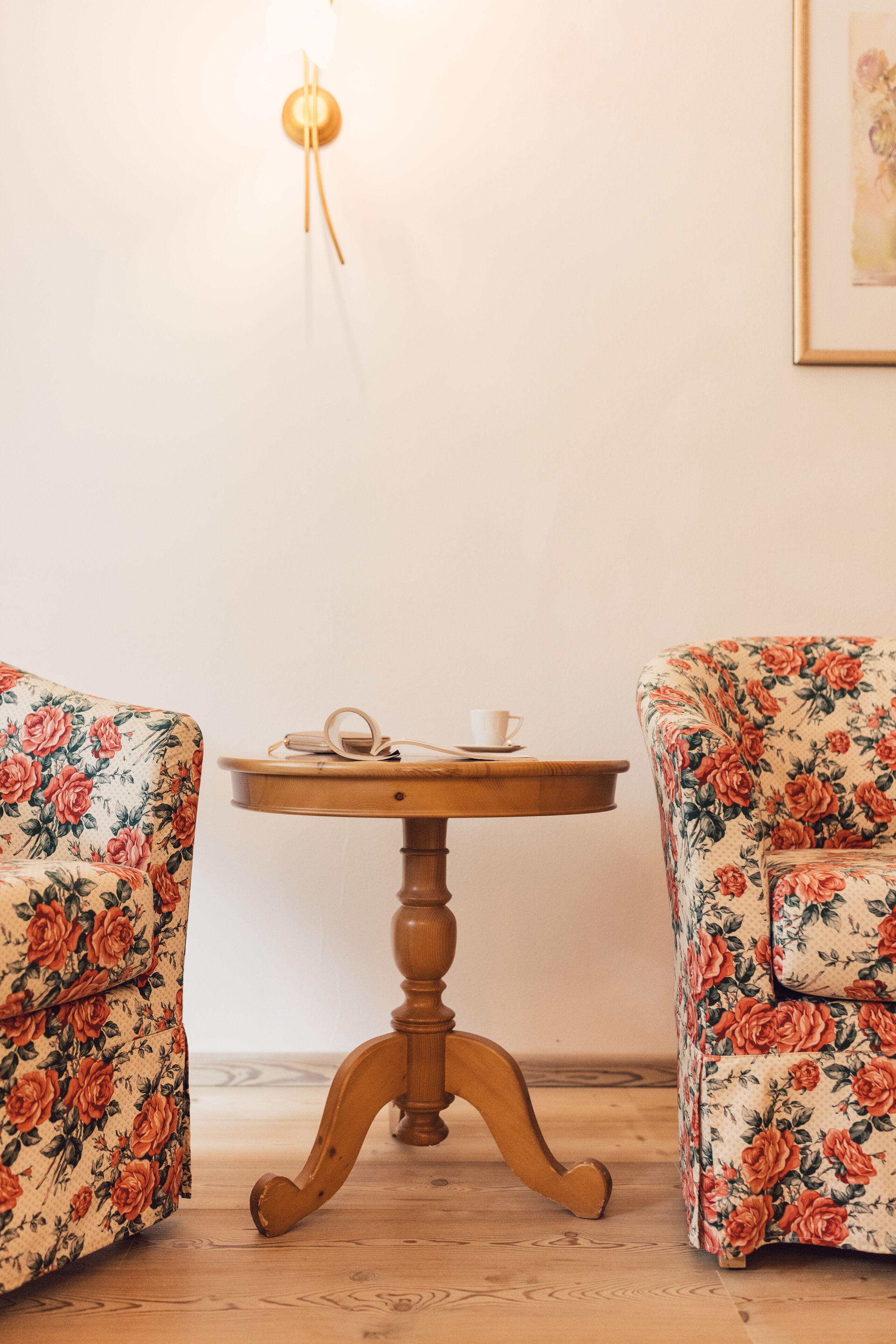A room with two floral armchairs, a wooden table with a cup and saucer, and a wall-mounted light fixture.