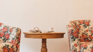 A room with two floral armchairs, a wooden table with a cup and saucer, and a wall-mounted light fixture.