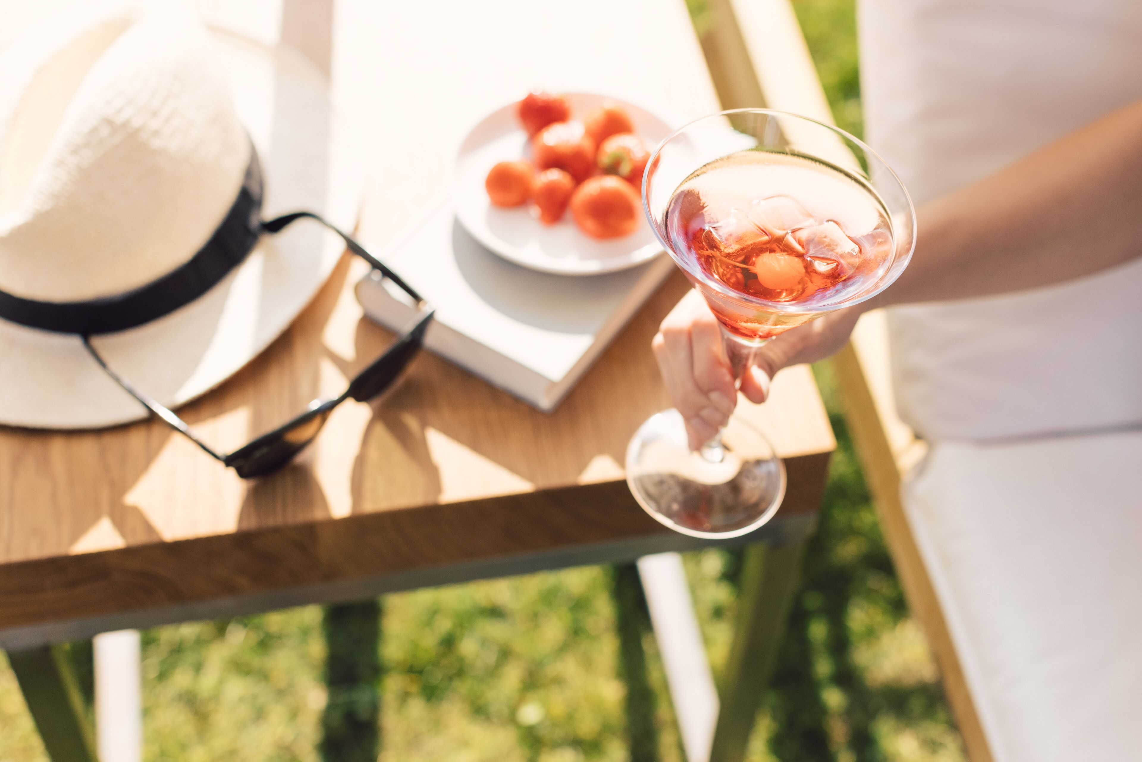 Une personne tenant un verre à cocktail rempli, une assiette de fruits, un chapeau et des lunettes de soleil, le tout posé sur une table en bois à l'extérieur.