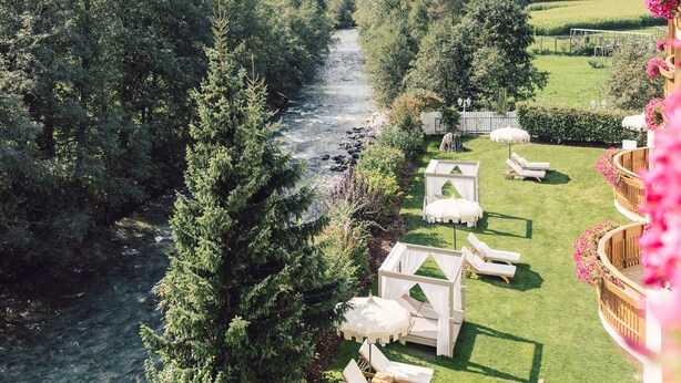 Une rivière traverse un paysage verdoyant et luxuriant, avec des montagnes en toile de fond. On y trouve des espaces de détente en plein air, avec des meubles blancs, et une personne qui se promène sur l'herbe.