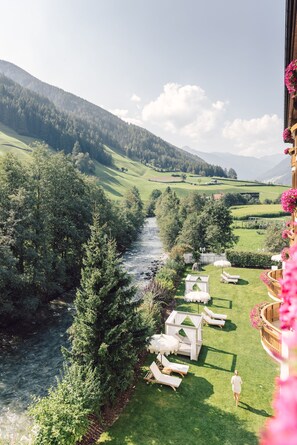 Un río serpentea por un paisaje verde y exuberante, con montañas al fondo. Hay zonas de estar al aire libre con mobiliario blanco y una persona caminando sobre el césped.