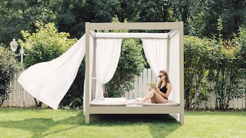 Relax in a serene outdoor setting with a four-poster bed surrounded by lush greenery and a white fence.