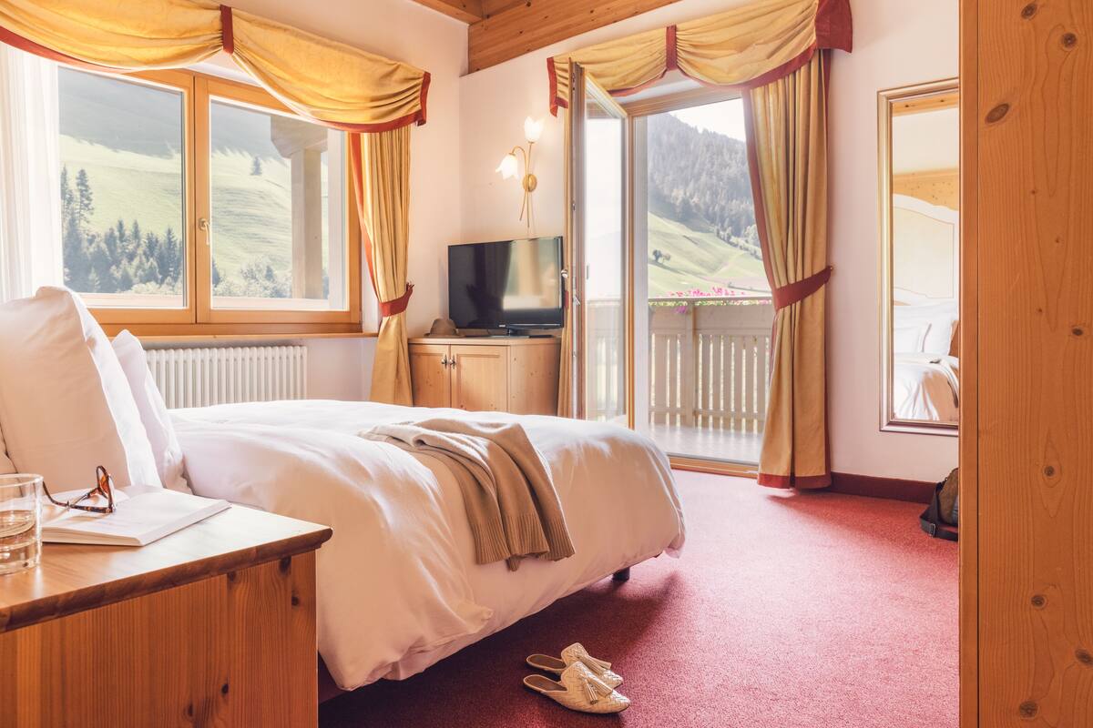 Een slaapkamer met een bed, een televisie, een houten bureau en uitzicht op een balkon met gordijnen.