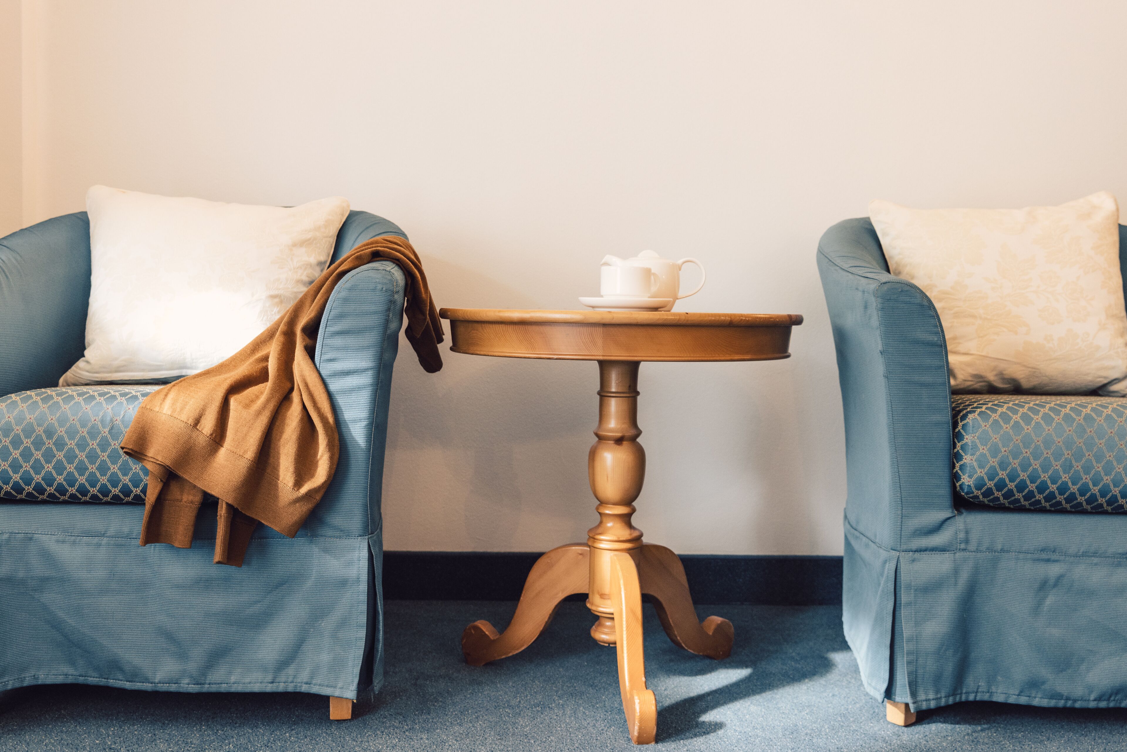 Deux fauteuils bleus ornés de coussins à motifs, une table en bois sur laquelle se trouvent une tasse et une soucoupe blanches, et un plaid marron drapé sur l’un des fauteuils.